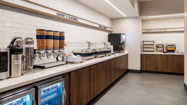 A breakfast bar with a coffee machine, a beverage cooler, and containers of whole milk, soy milk, and almond milk. - Tru By Hilton Atlanta Airport College Park (College Park)