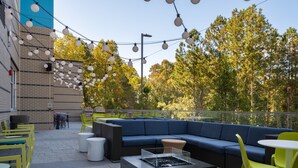 An outdoor patio with a black fire pit, a blue sofa, yellow and green chairs, and string lights. - Tru By Hilton Atlanta Airport College Park (College Park)