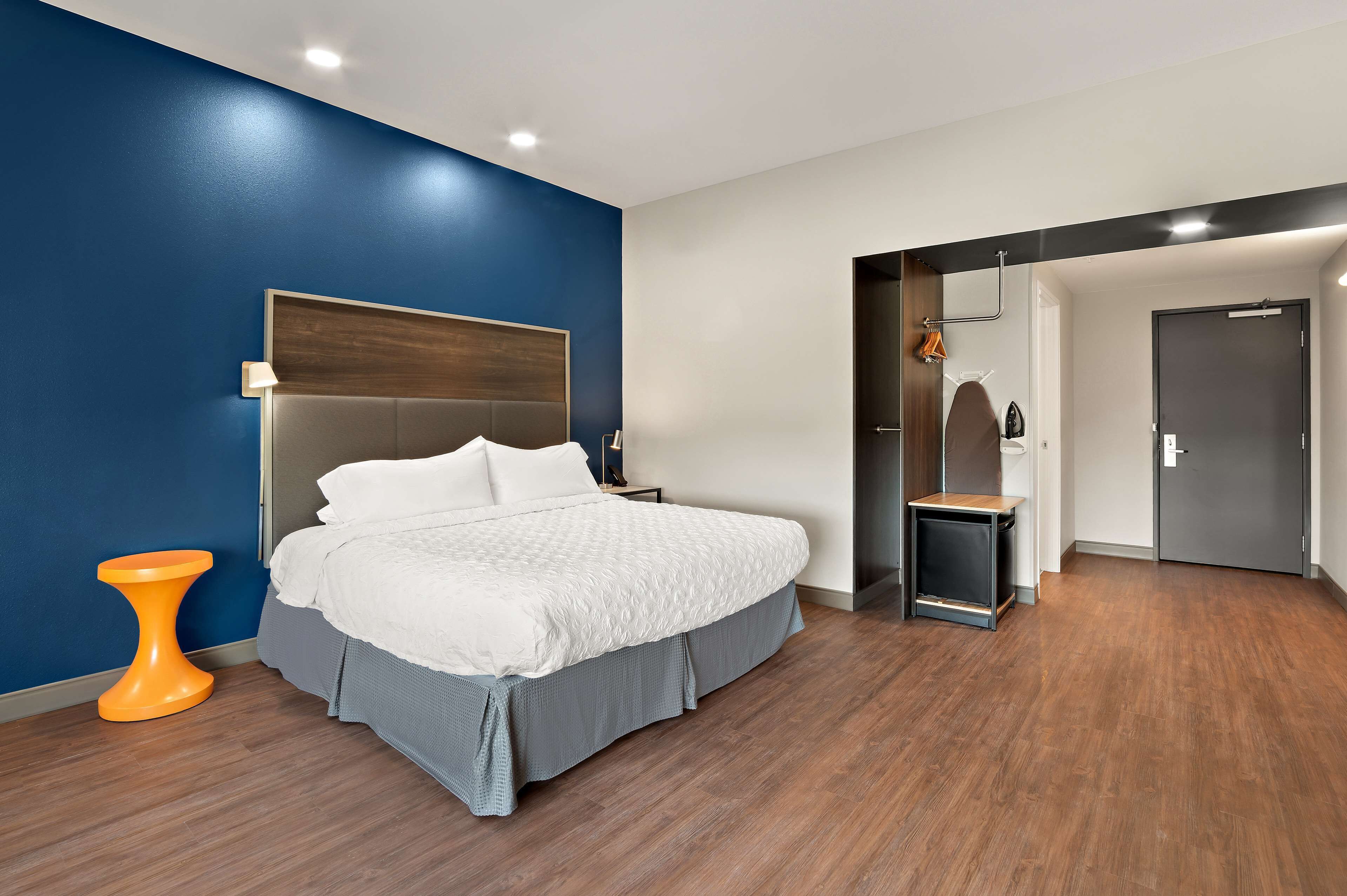 Una habitación de hotel moderna con una cama grande, un cabecero de madera, una mesita de noche naranja y un banco gris.