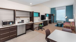 Una habitación de hotel moderna con kitchenette, un escritorio con computadora, televisión, sofá y ventana con vistas.