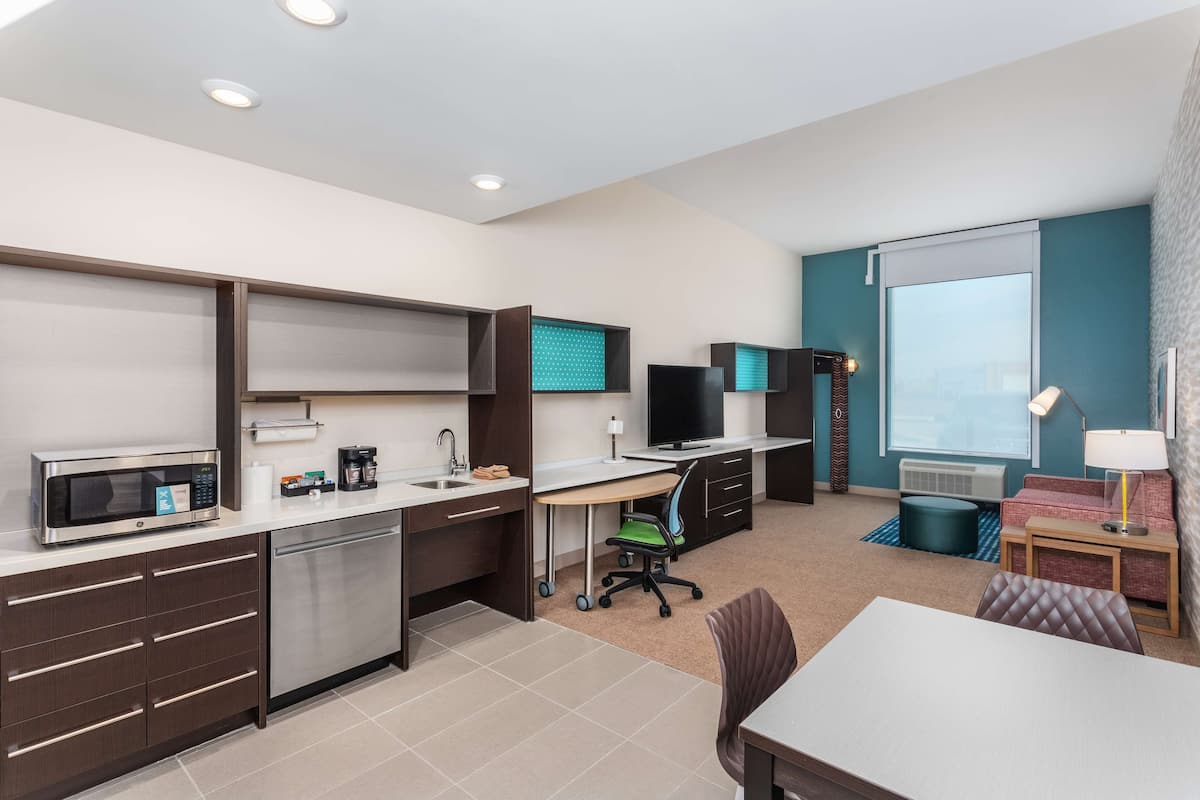 Una habitación de hotel moderna con kitchenette, un escritorio con computadora, televisión, sofá y ventana con vistas.