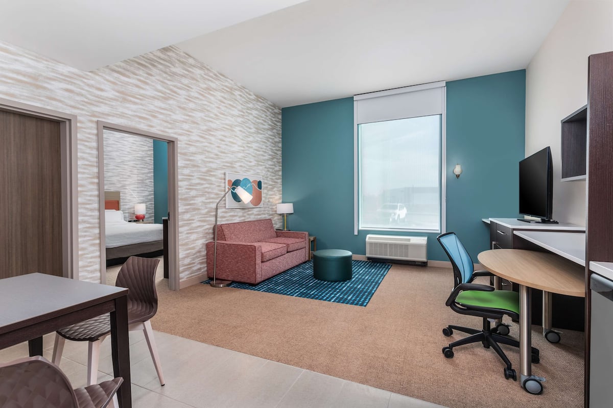 Una habitación de hotel moderna con kitchenette, un sofá, un escritorio y un ventanal grande.