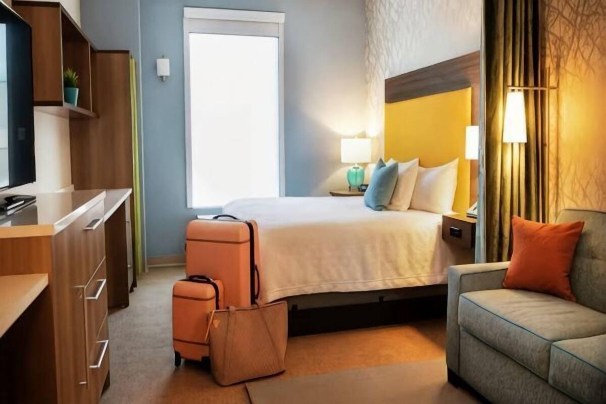 A hotel room with a bed, a desk, a suitcase, a sofa, and a nightstand with lamps.