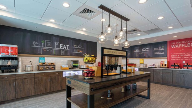 A modern cafe interior with a central wooden table, hanging light fixtures, and a chalkboard wall with menu items. - Hampton Inn Marianna I-10 (Marianna)