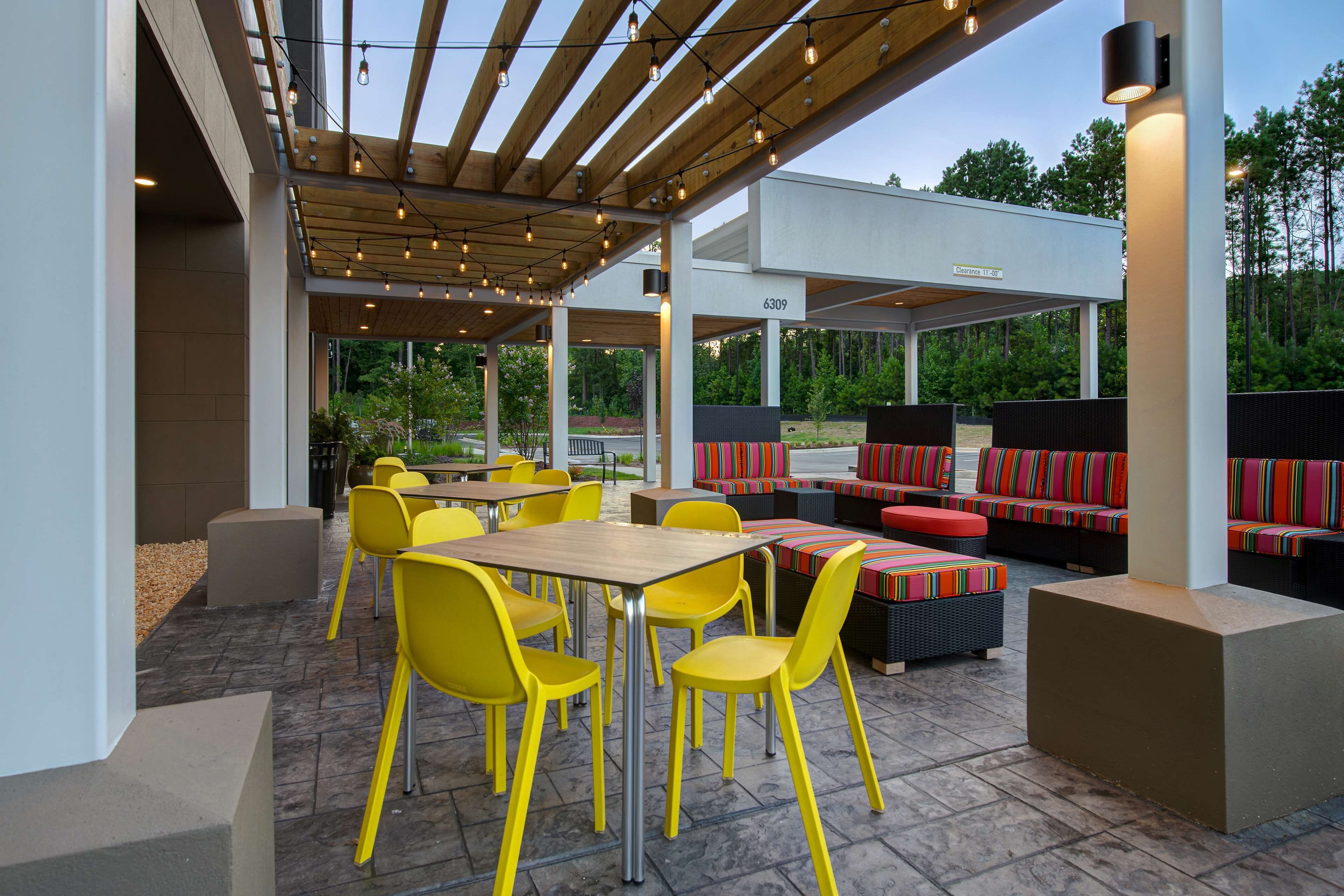 An outdoor patio area with yellow chairs and tables, a covered seating area with colorful cushions, and a view of trees in the background.