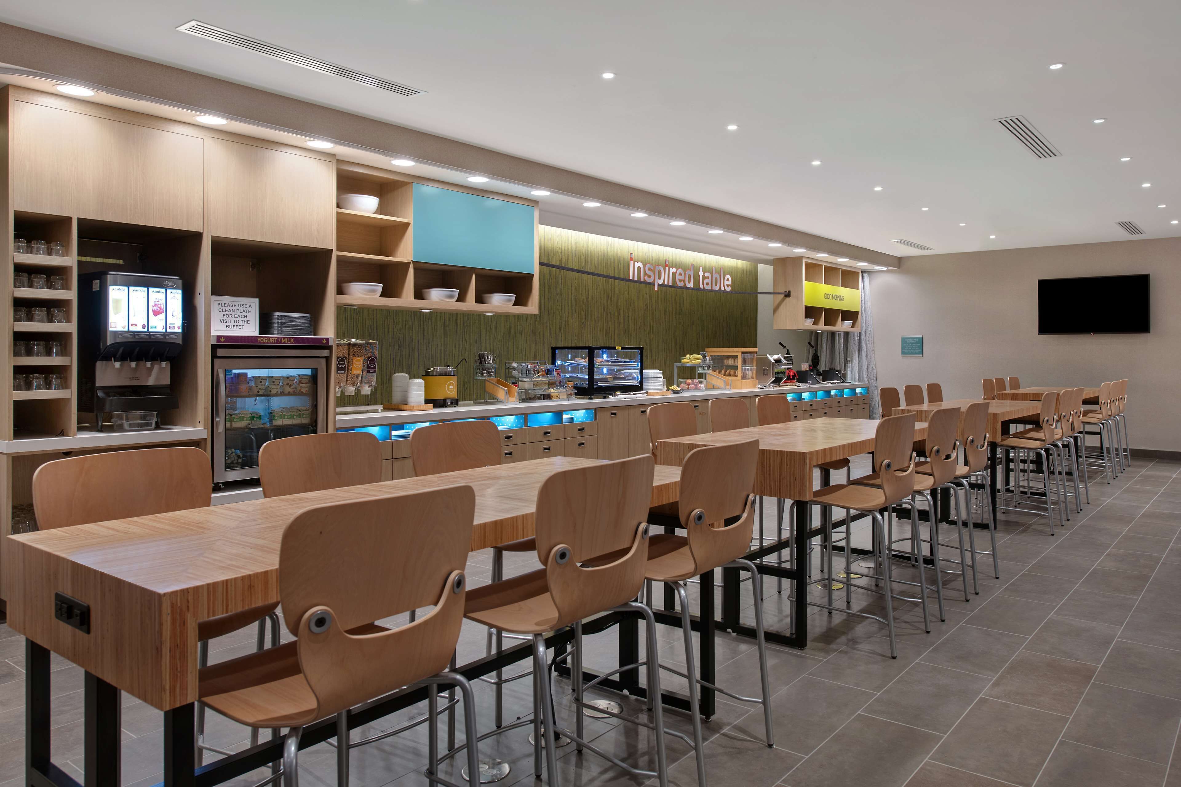 A modern restaurant with long wooden tables and chairs, a self-service beverage station, and a counter with a digital menu display.
