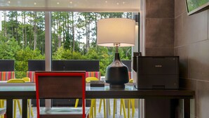 Una habitación moderna con escritorio, lámpara e impresora Brother. Hay sillas con cojines de colores y vistas a árboles a través de amplias ventanas.