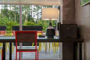Una habitación moderna con escritorio, lámpara e impresora Brother. Hay sillas con cojines de colores y vistas a árboles a través de amplias ventanas.