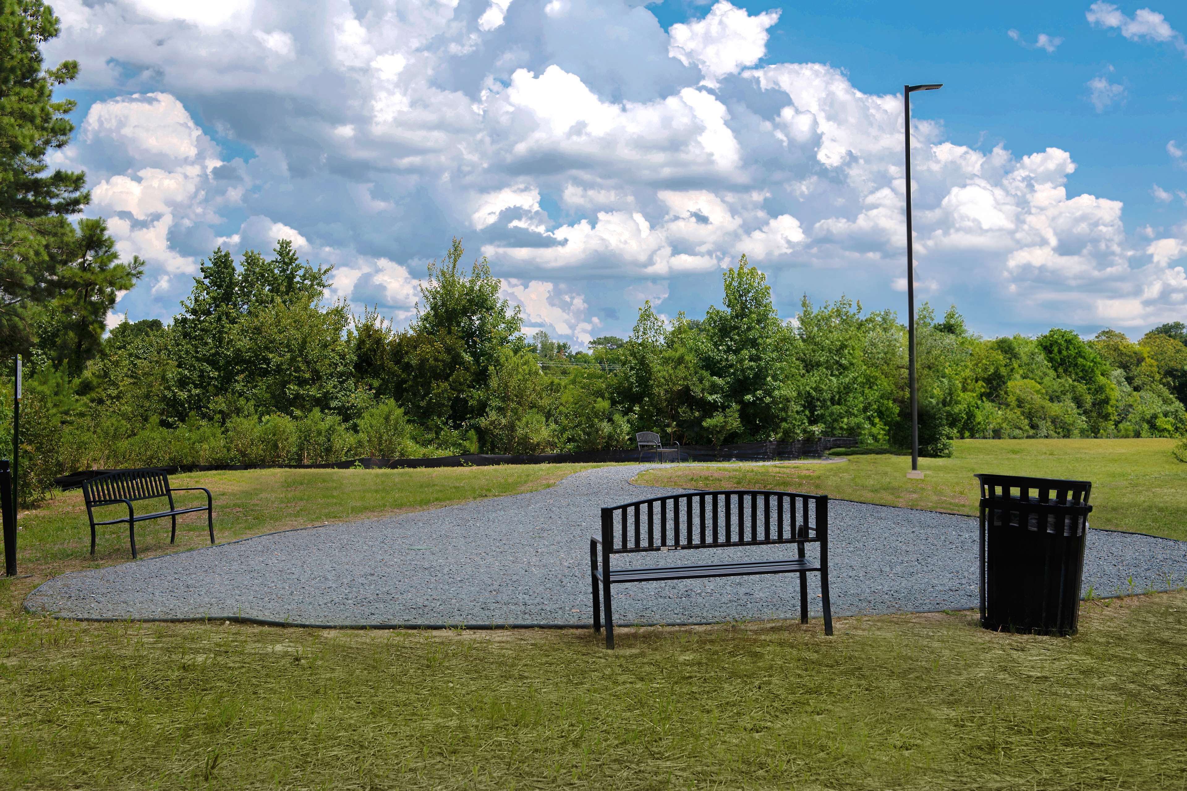 A park with a gravel path, benches, and a trash can.