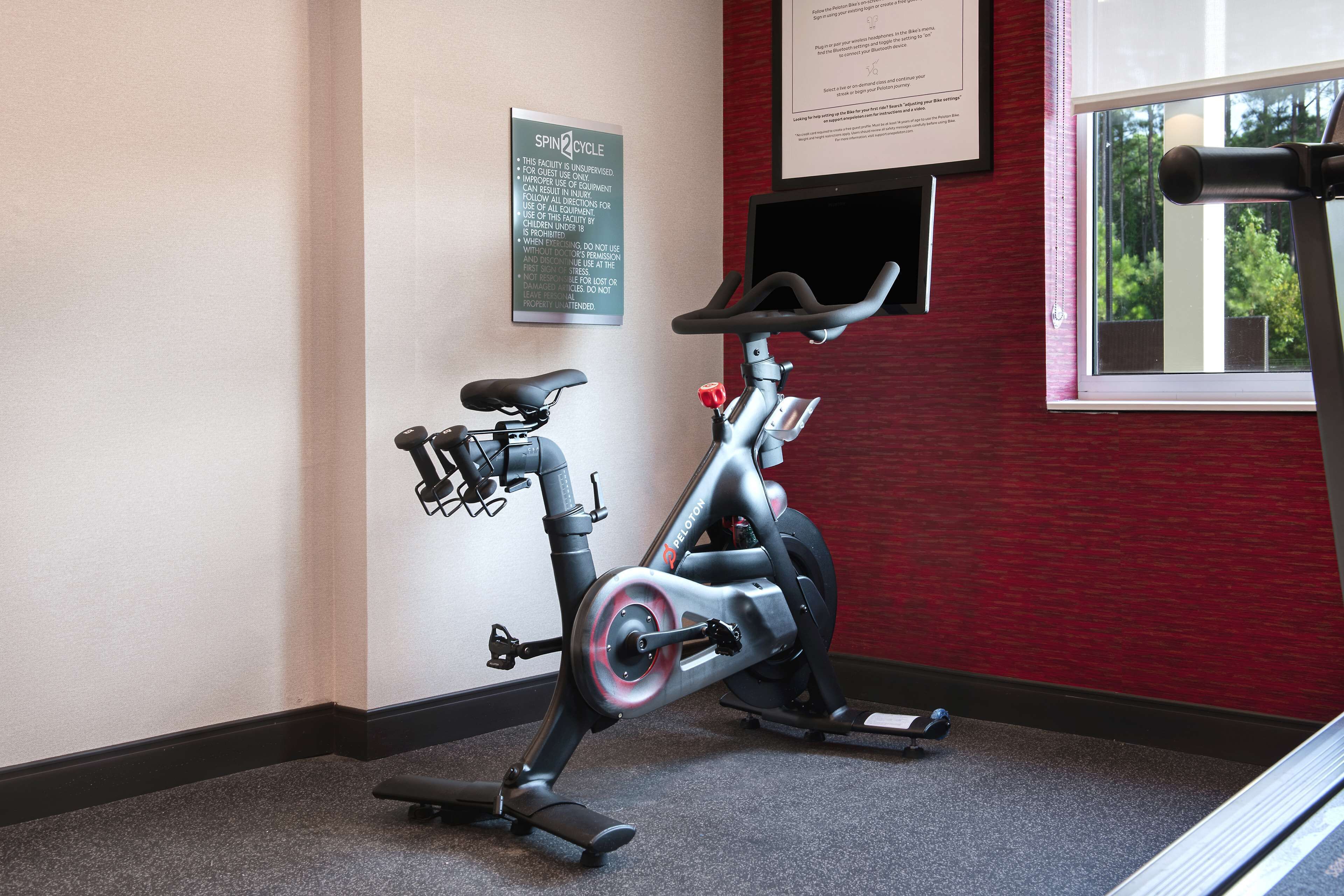 A stationary bike in a room with a red wall, a framed sign on the wall, and a window with a view of trees.