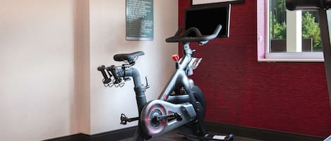 A stationary bike in a room with a red wall, a framed sign on the wall, and a window with a view of trees.