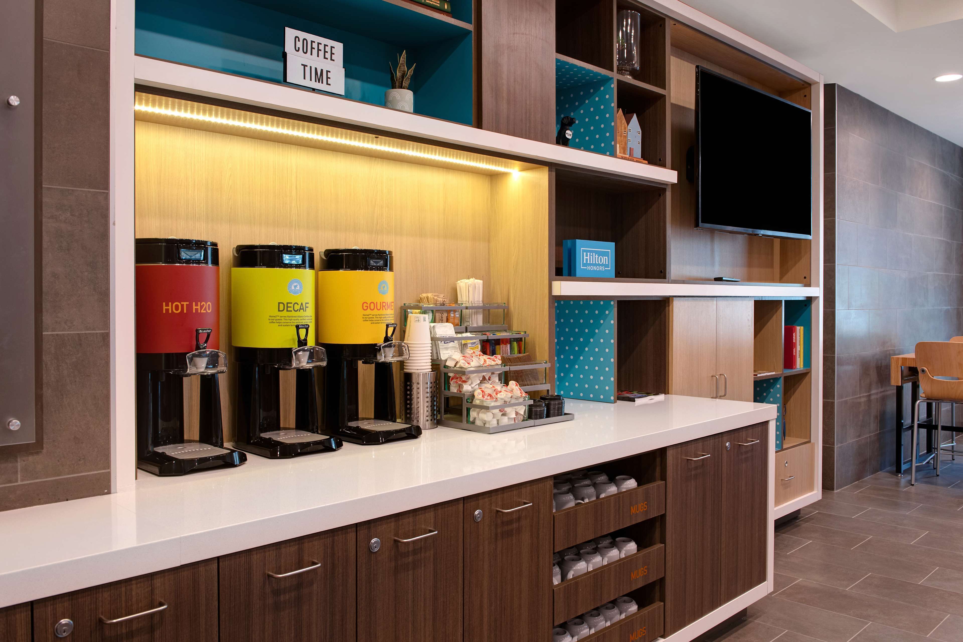 A hotel reception area with a coffee station, a flat-screen TV, and a seating area.