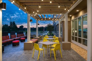 An outdoor patio area with yellow chairs and tables, a covered seating area with red and black cushions, and a view of the sky and trees in the background. - Home2 Suites By Hilton Fayetteville North (Fayetteville)