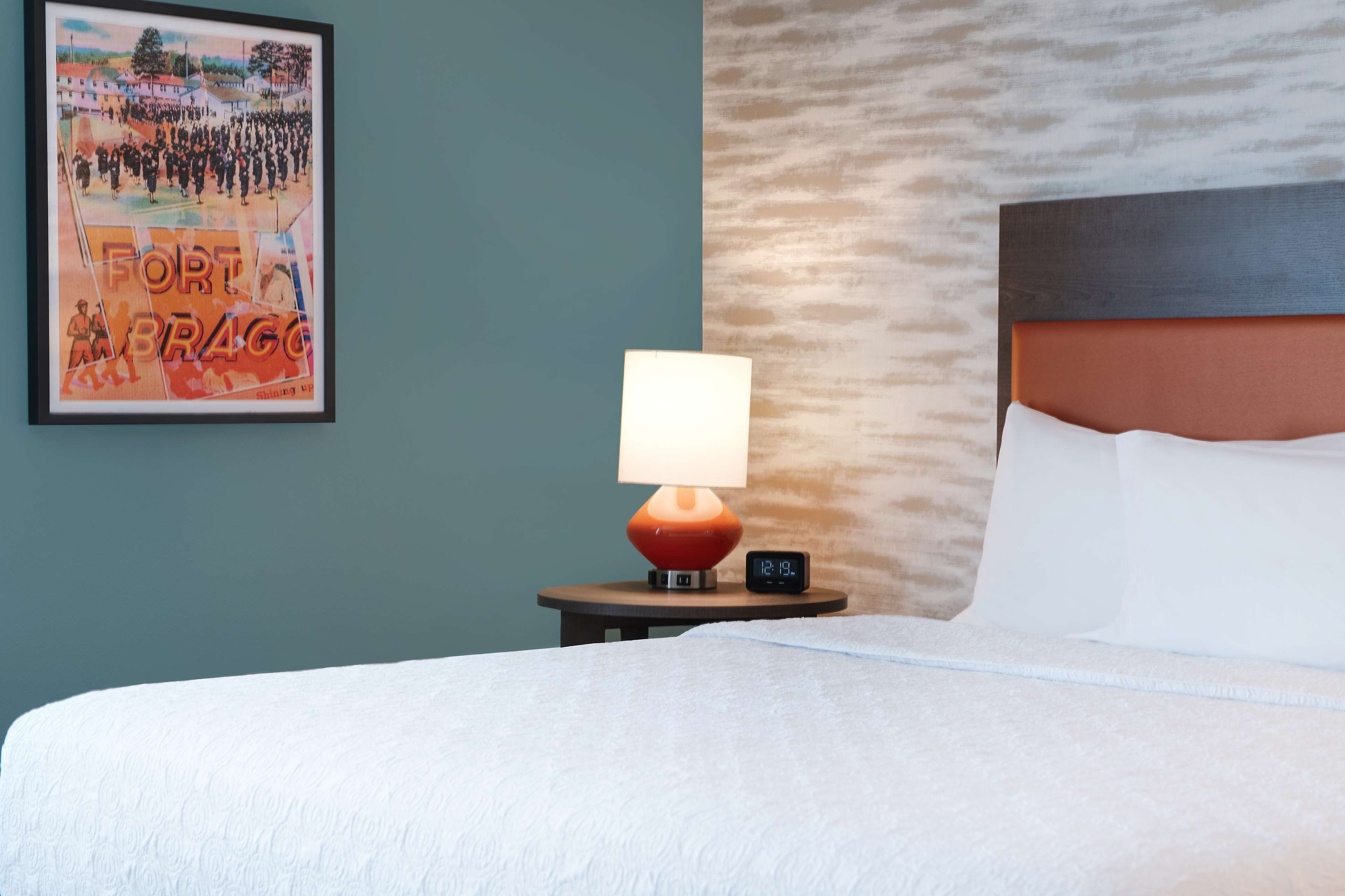 A hotel room with a bed, a nightstand with a lamp and a digital clock, and a framed picture on the wall.