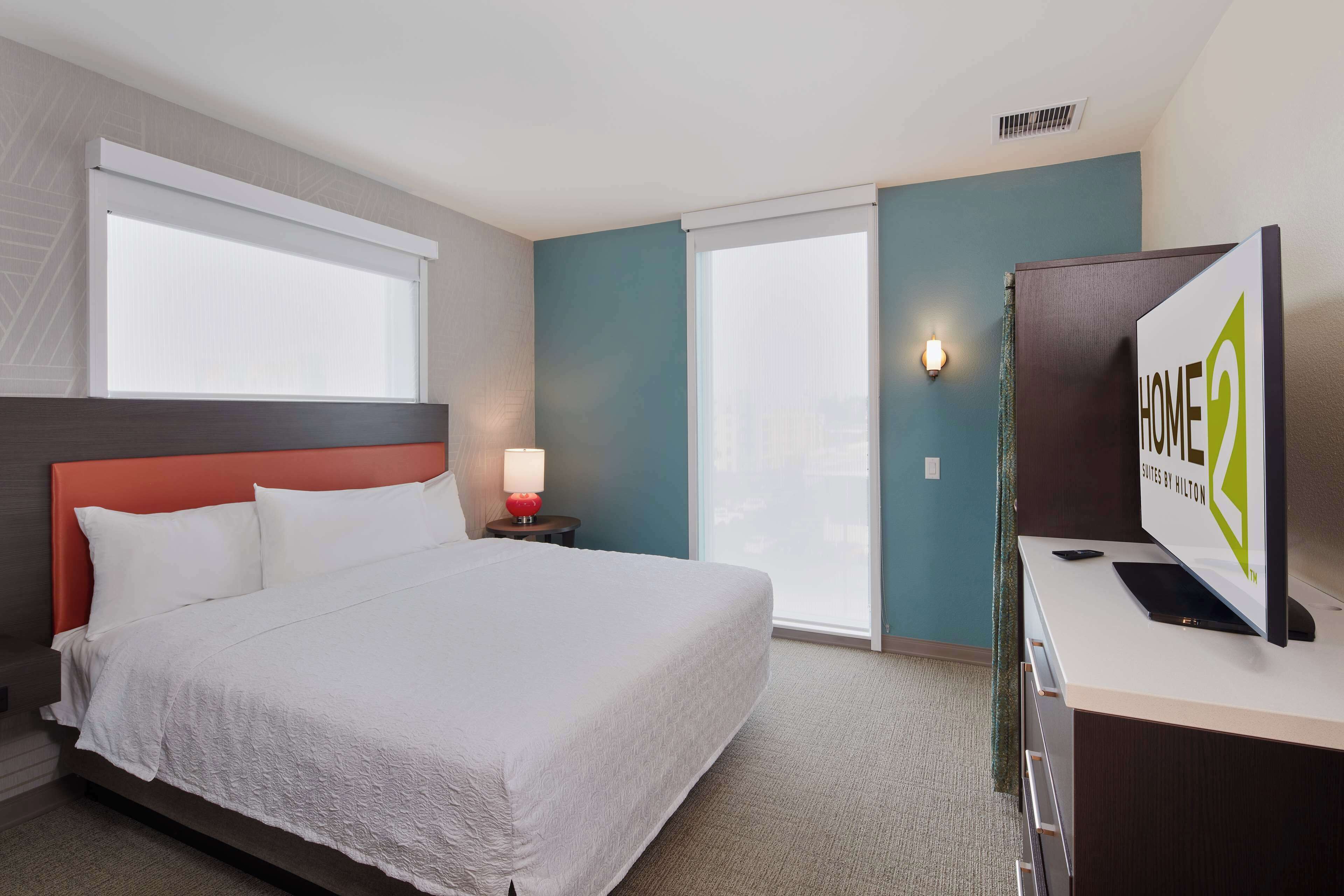 A hotel room with a bed, a desk, a TV, and a window with blinds.