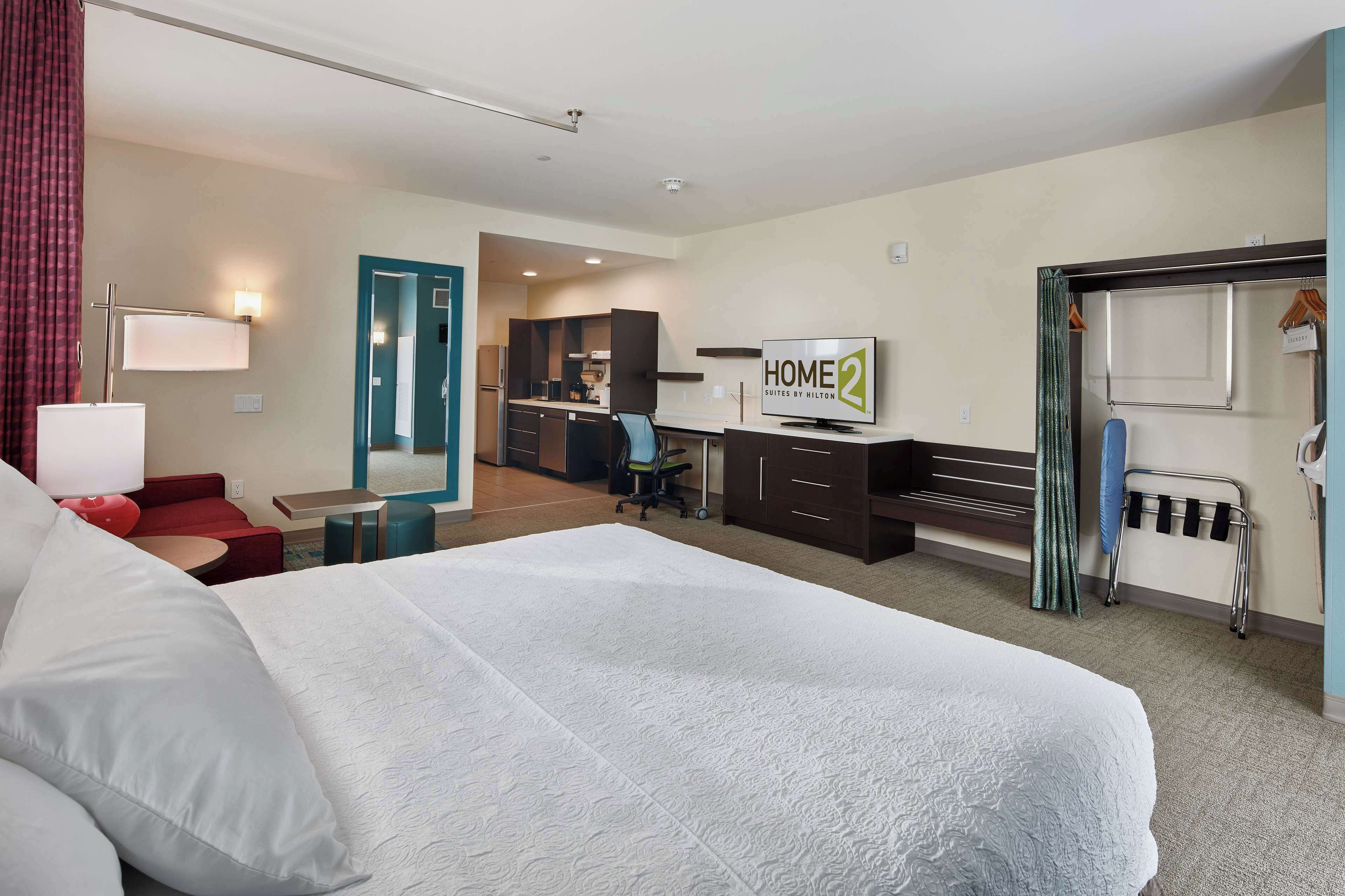 A hotel room with a large bed, a desk with a computer, a TV, and a kitchenette in the background.