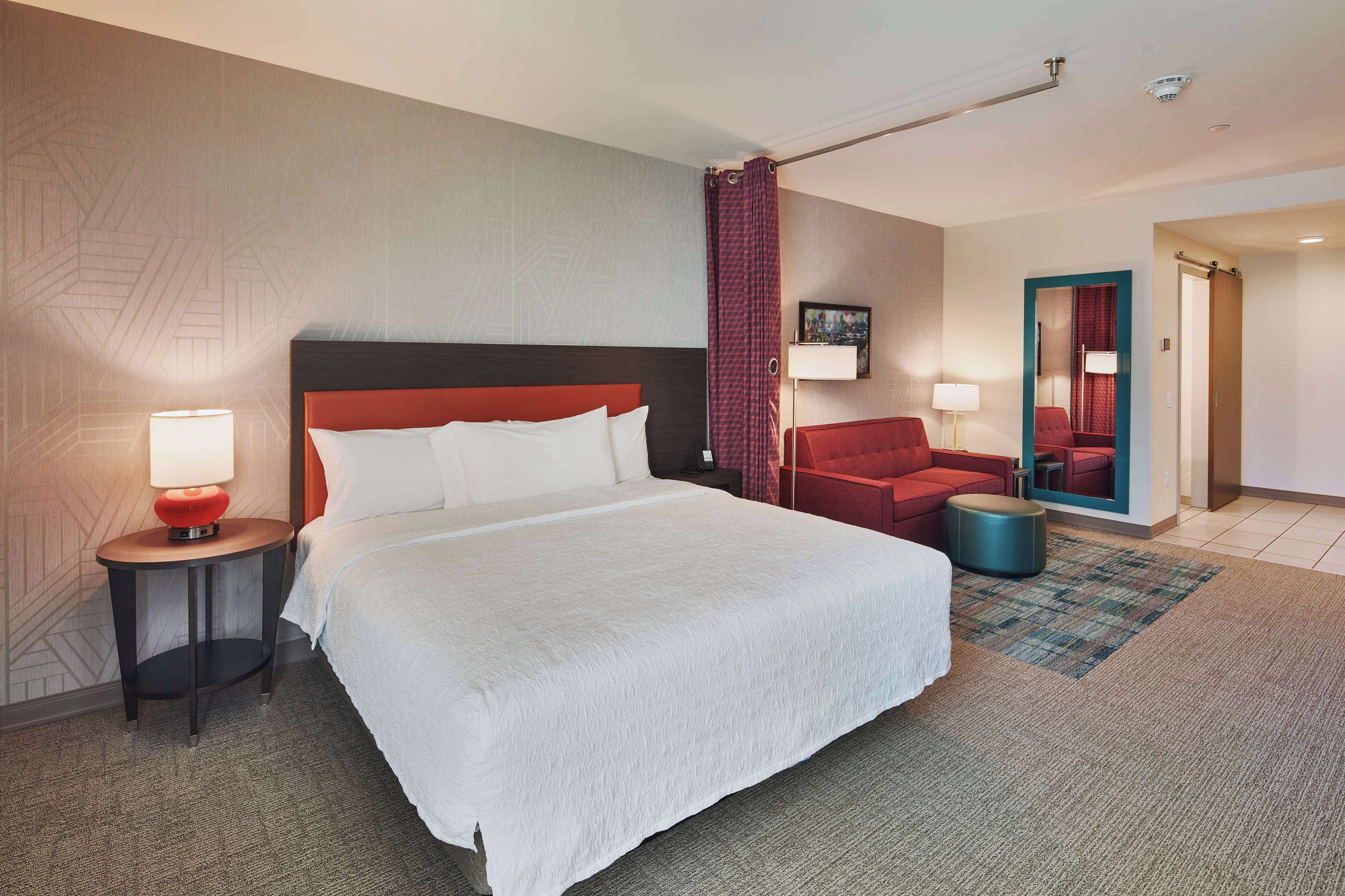 A hotel room with a large bed, a red chair, a small table with a lamp, and a mirror on the wall.