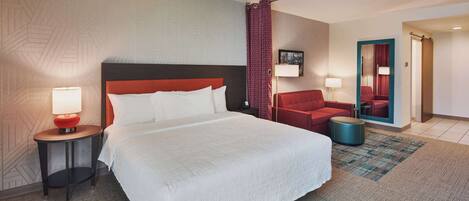 A hotel room with a large bed, a red chair, a small table with a lamp, and a mirror on the wall.