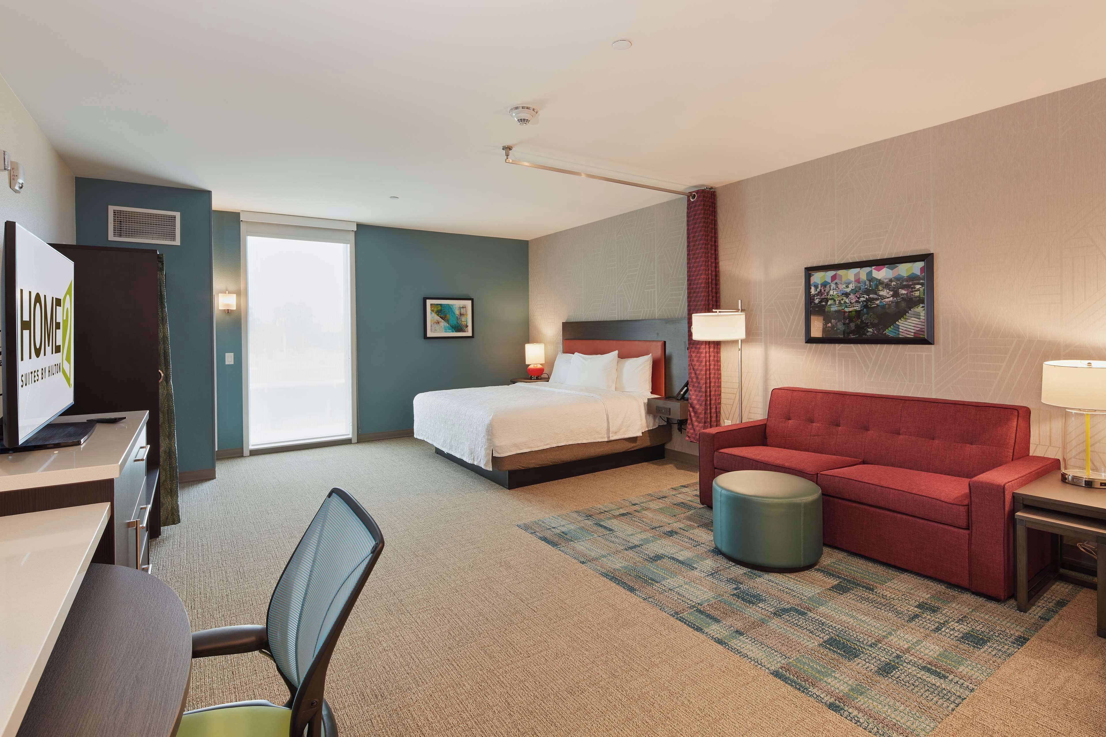 A hotel room with a bed, a red sofa, a desk with a computer, a chair, and a small round ottoman.