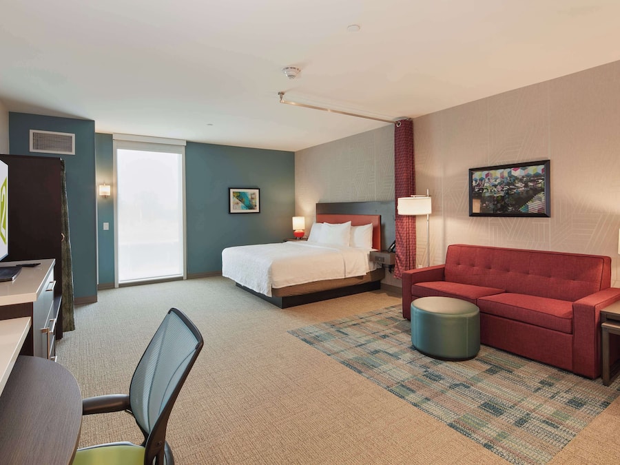 A hotel room with a bed, a red sofa, a desk with a computer, a chair, and a small round ottoman.