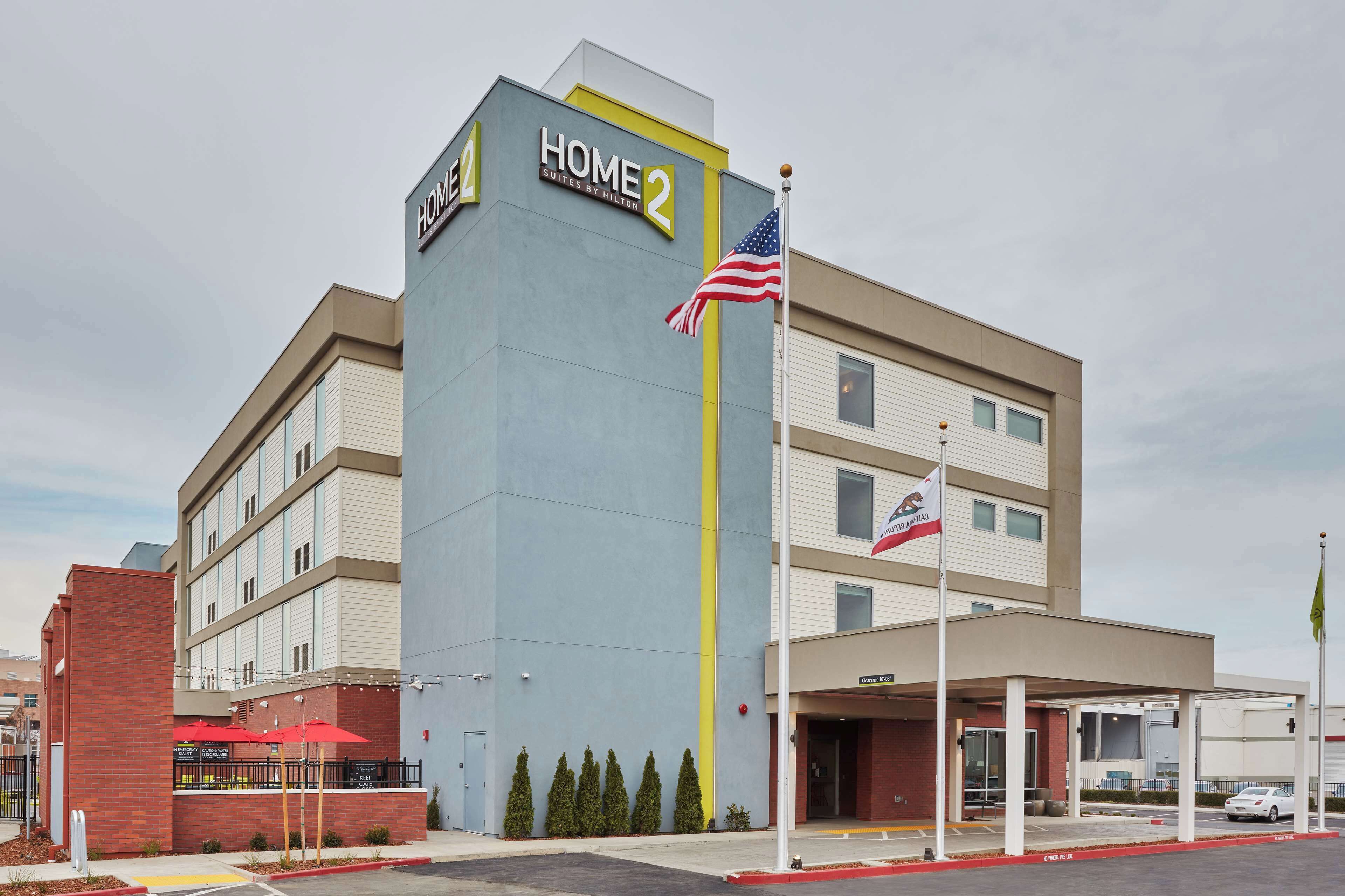 Un hotel llamado "Home 2 Suites by Hilton" con un diseño moderno, una entrada cubierta y un asta con la bandera estadounidense.