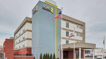 A hotel named "Home 2 Suites by Hilton" with a modern design, a covered entrance, and a flagpole with an American flag.