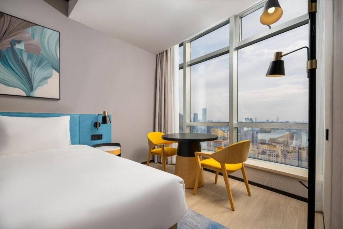 A hotel room with a bed, a round table with chairs, a large window with a city view, and a painting on the wall.