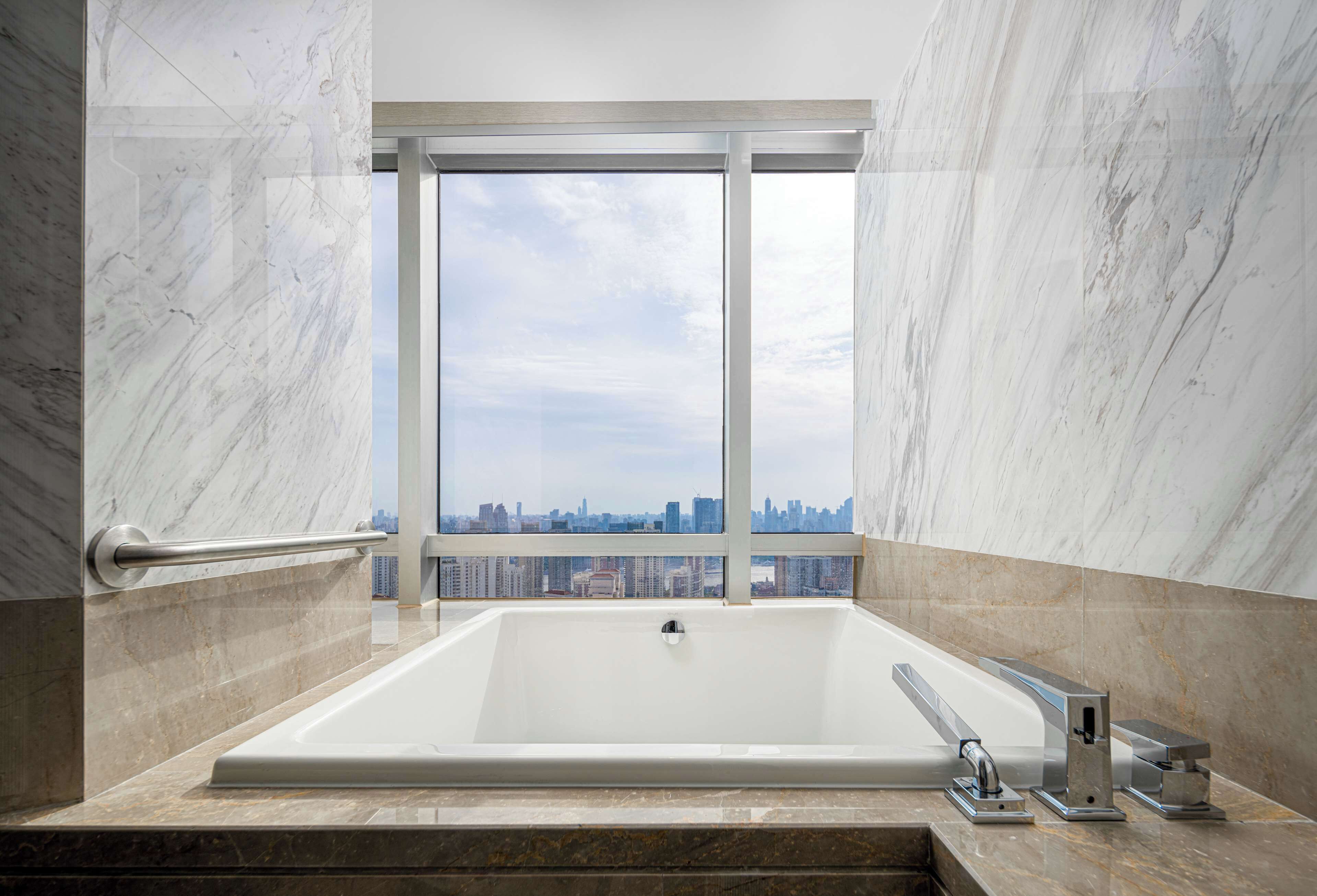 Ein modernes Badezimmer mit einer großen Badewanne, Marmorwänden und Blick auf die Stadt durch ein großes Fenster.