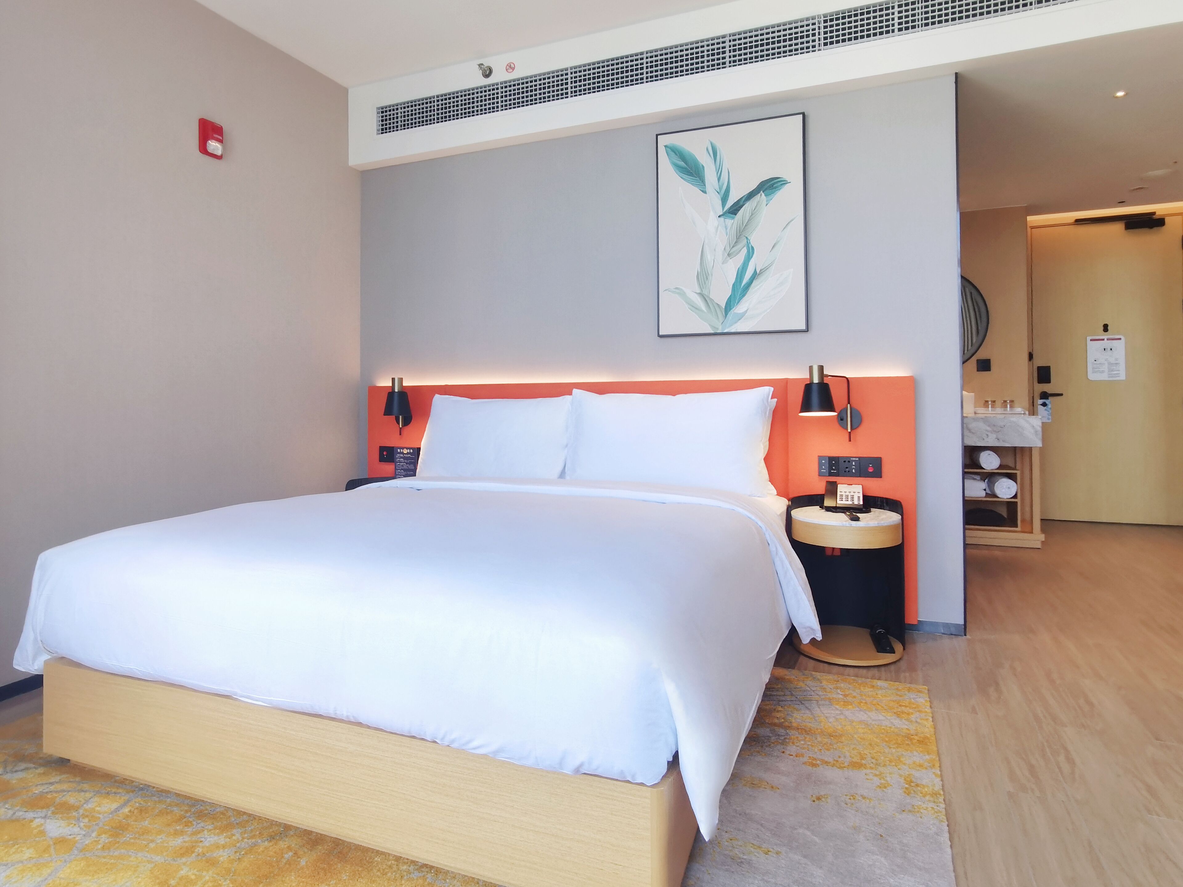 Habitación de hotel con una cama grande, cabecera con iluminación integrada, mesita de noche con teléfono y lámpara, y un mueble de madera pequeño con toallas.
