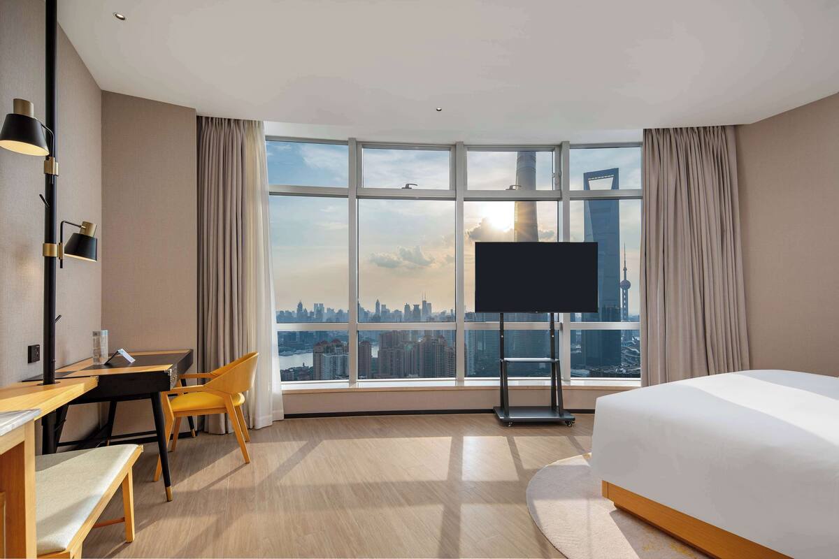 A hotel room with a large window, a bed, a desk with a chair, and a flat-screen TV.