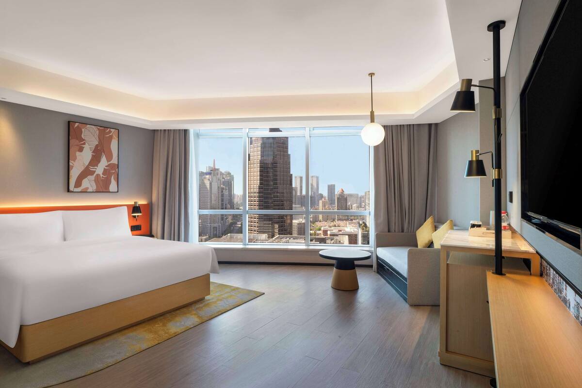 A modern hotel room with a large bed, a sofa, a small table, and a TV. The room has a city view through large windows.