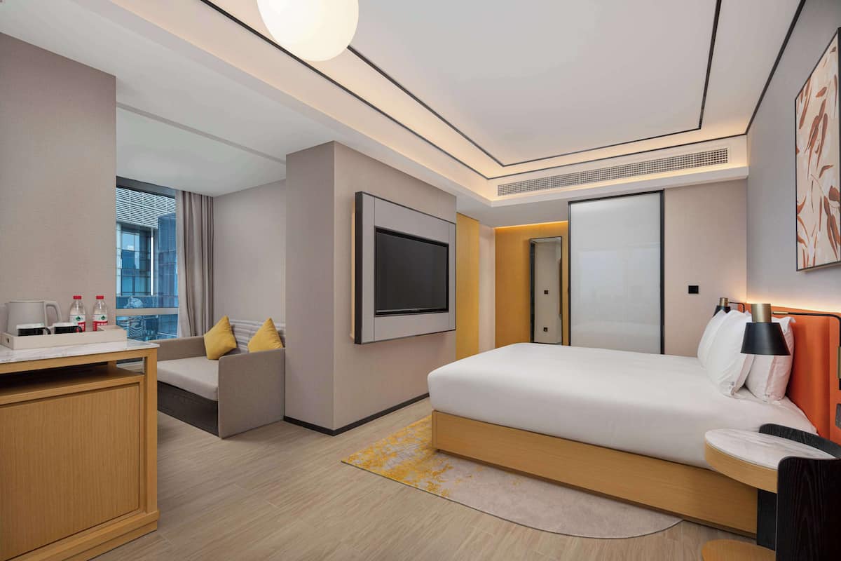 A modern hotel room with a bed, a desk, a sofa, a TV, and a window with curtains.