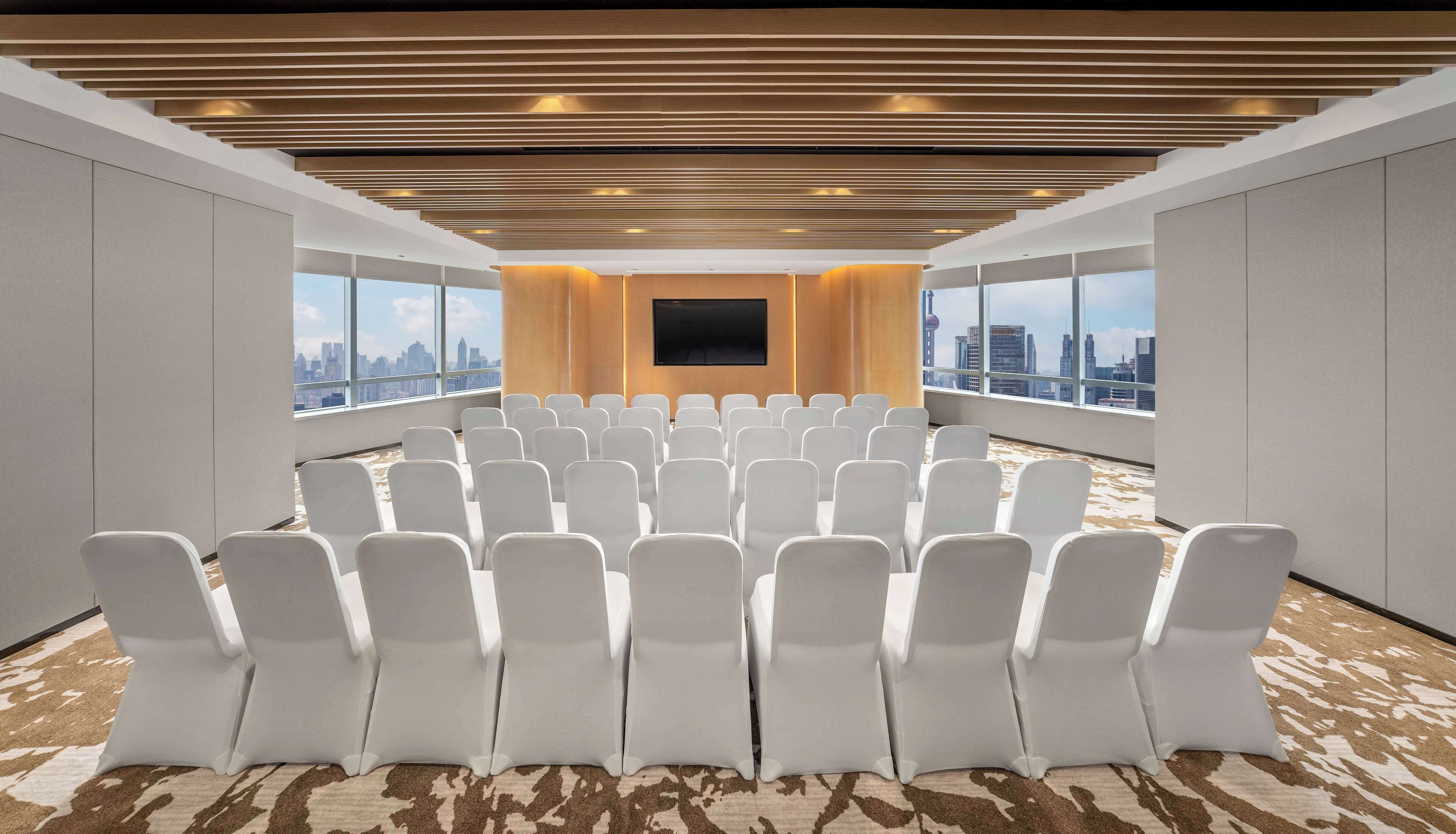 Un salón de conferencias con filas de sillas blancas, una pantalla grande y vistas de la ciudad a través de amplios ventanales.