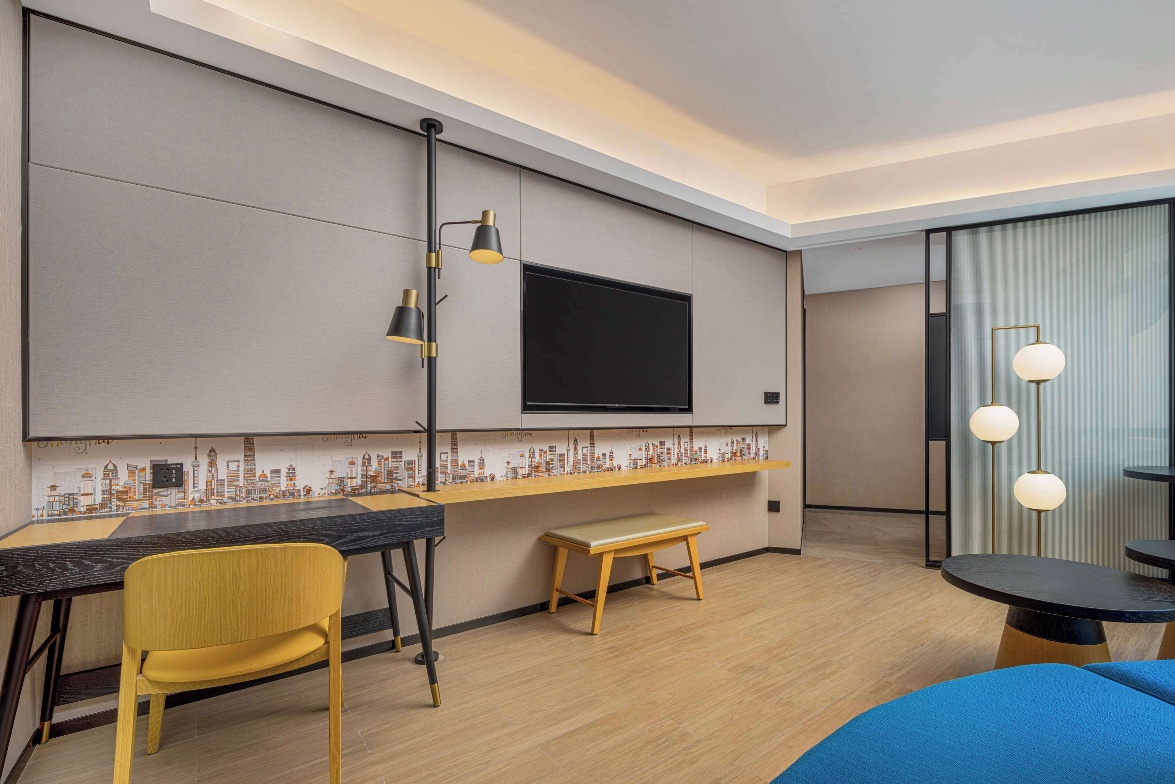 A modern hotel room with a flat-screen TV mounted on the wall, a wooden bench, a yellow chair, and a small table with a blue cushion.