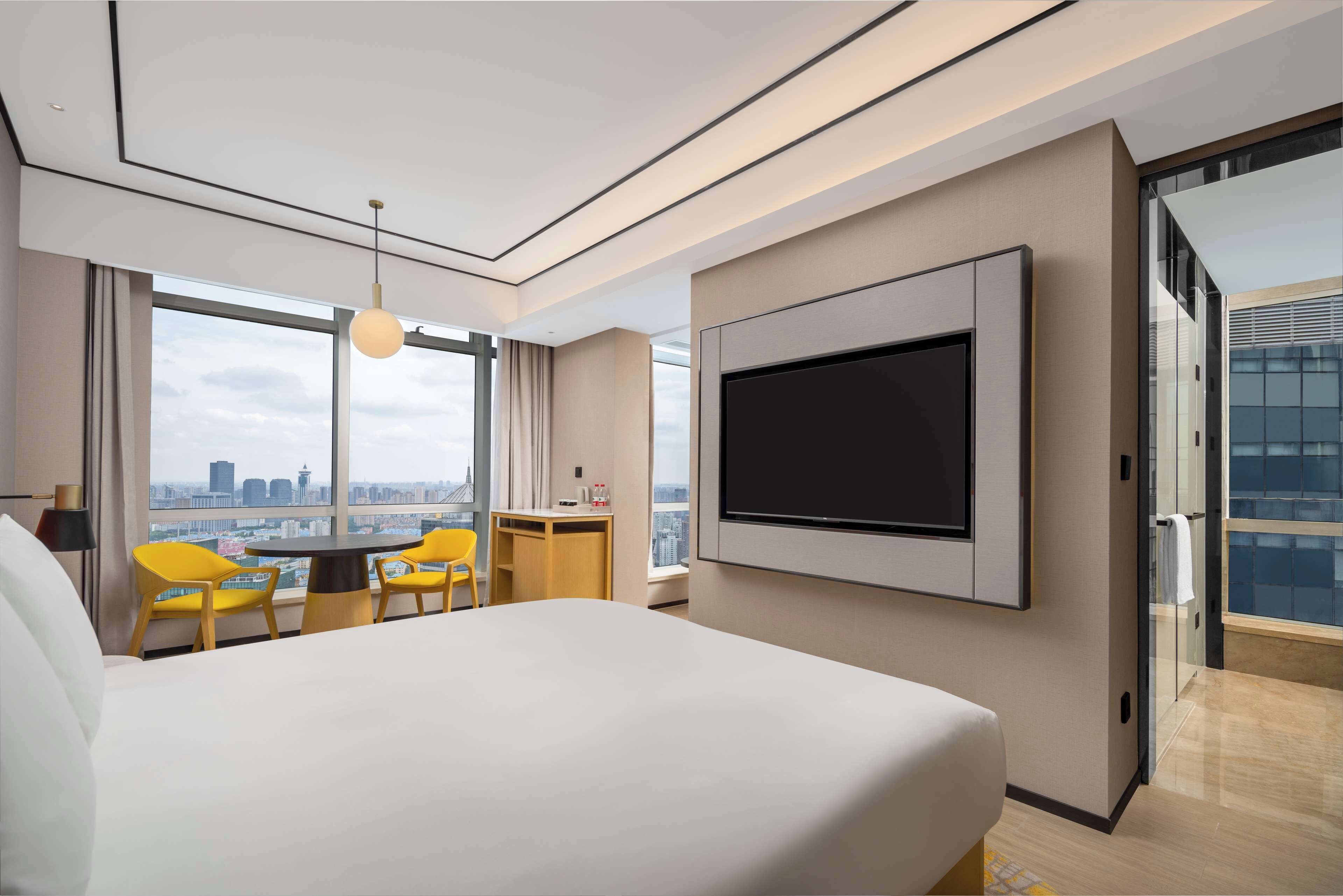 A modern hotel room with a large bed, a TV mounted on the wall, a desk with a chair, and a view of the city through large windows.