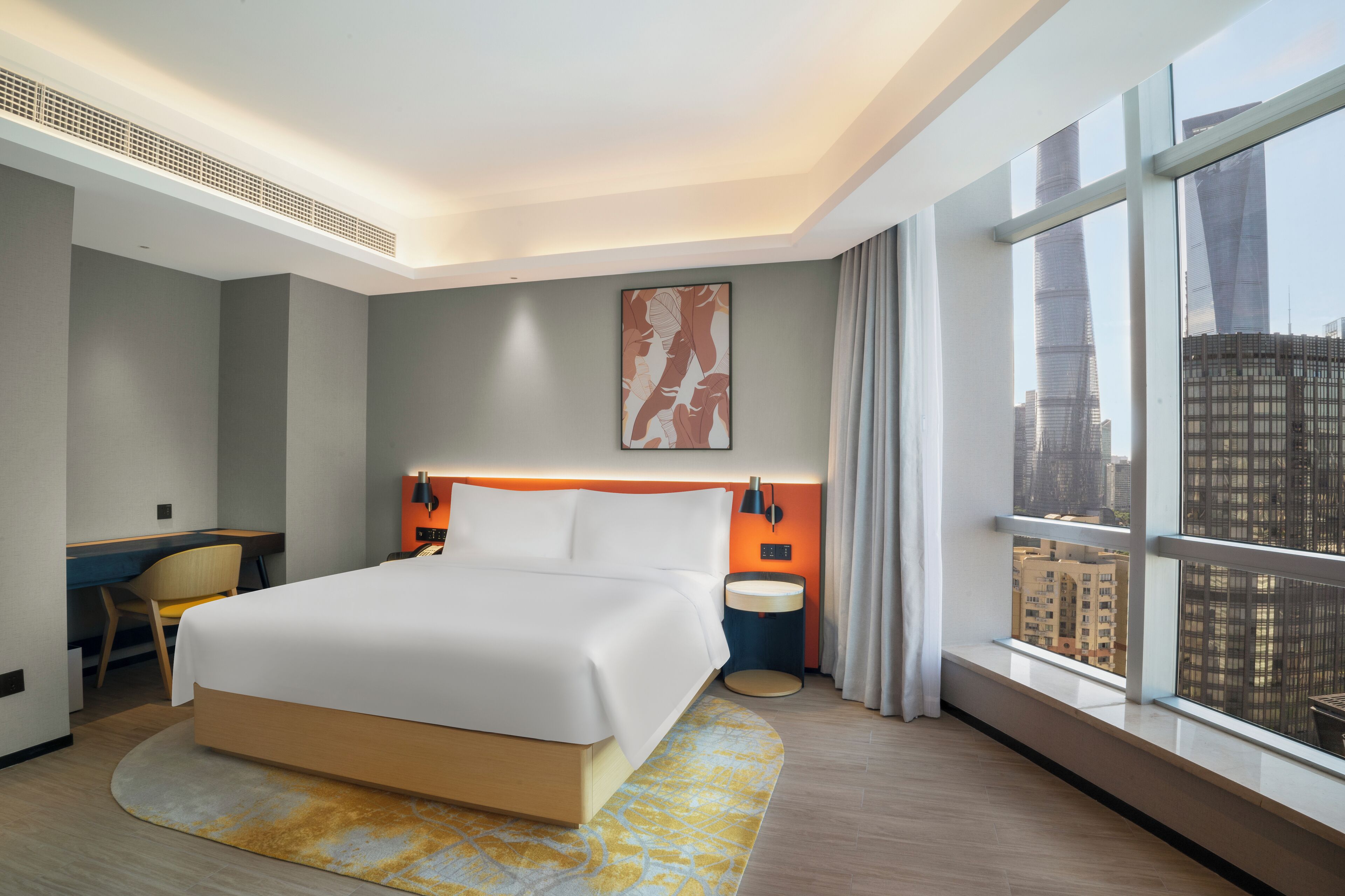 A modern hotel room with a large bed, a desk, and a view of the cityscape through large windows.