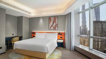 A modern hotel room with a large bed, a desk, and a view of the cityscape through large windows.