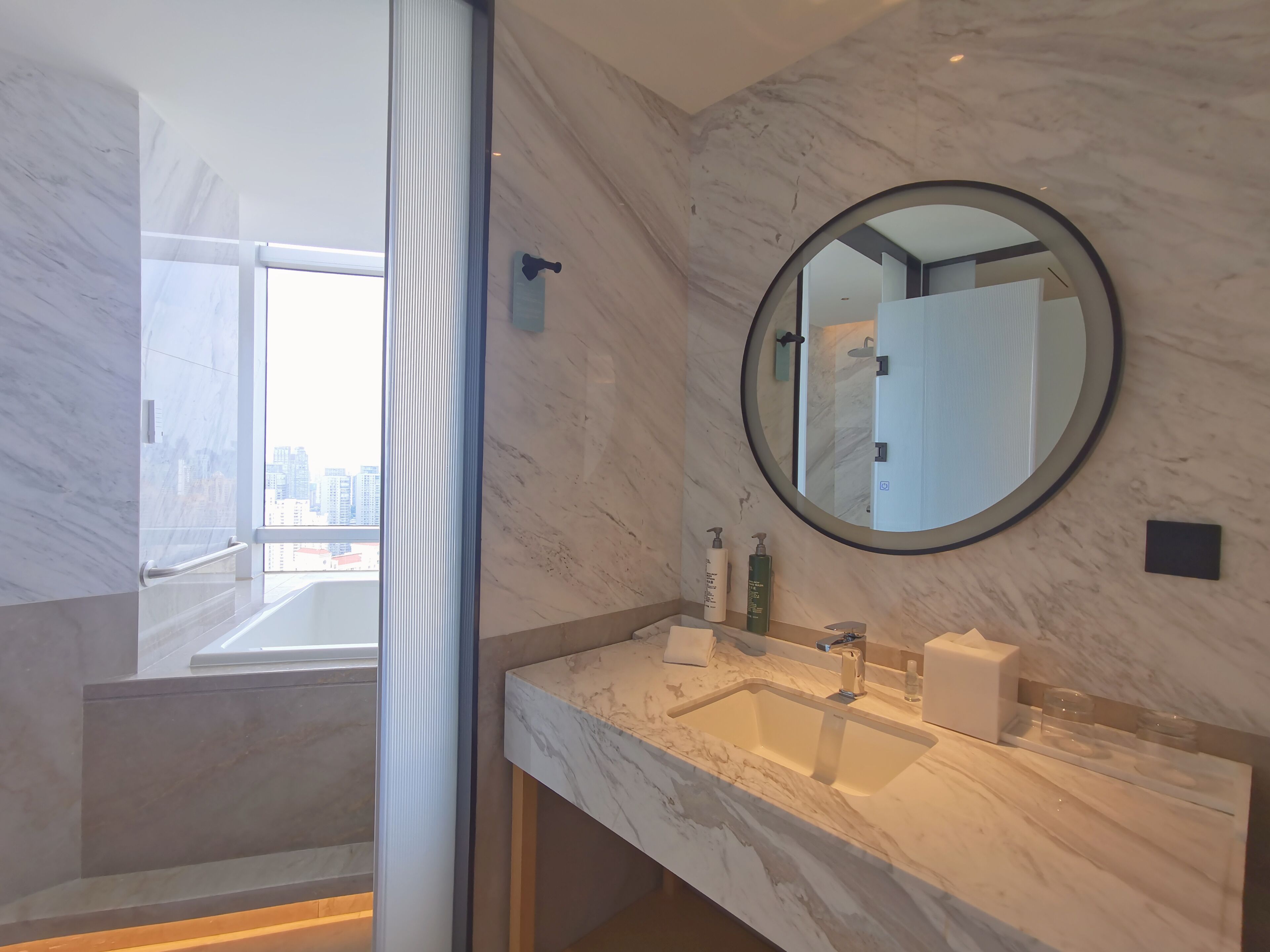 A modern bathroom with a marble sink, a round mirror, and a view of the city through a large window.