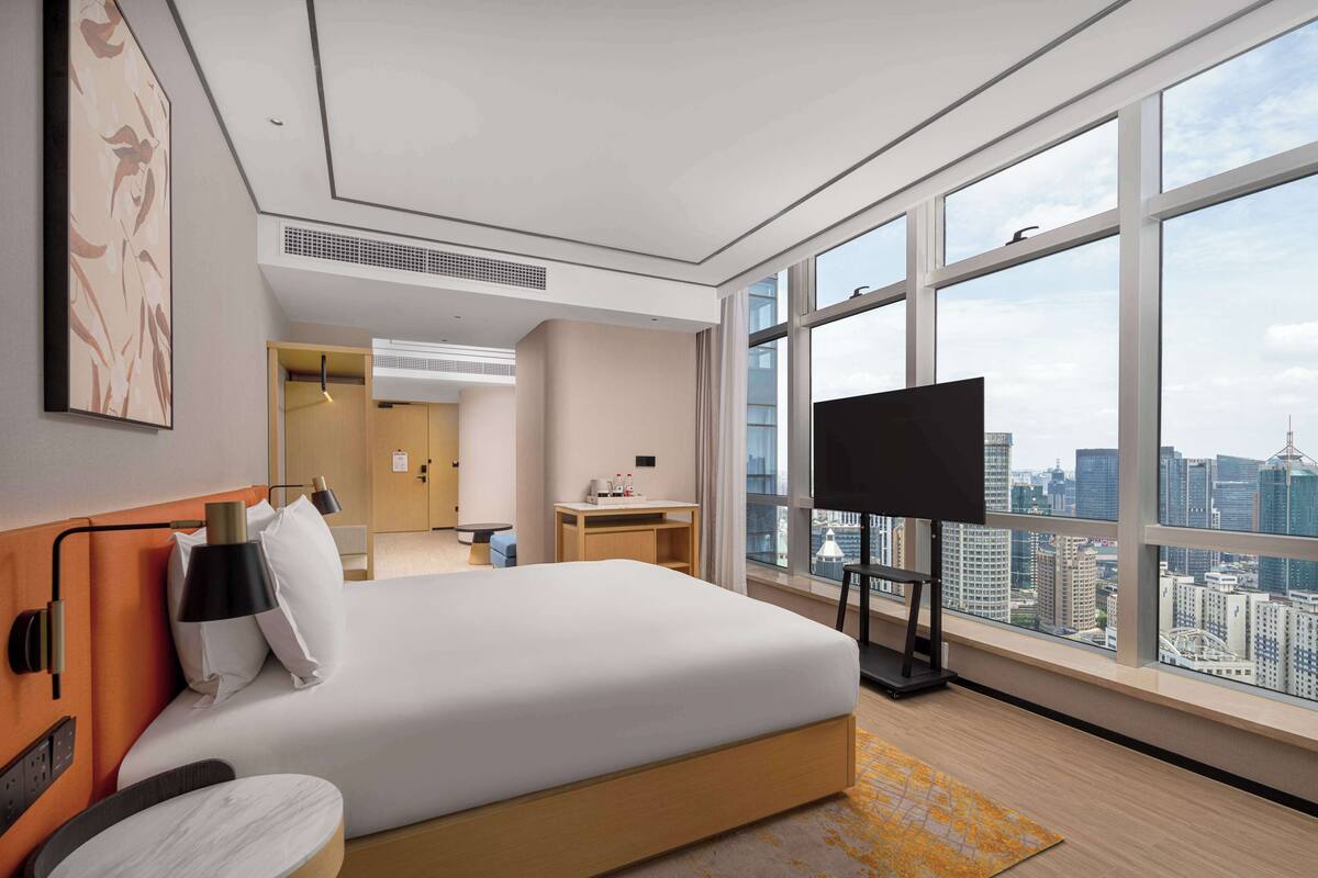 A modern hotel room with a large bed, a desk, a TV, and a city view.