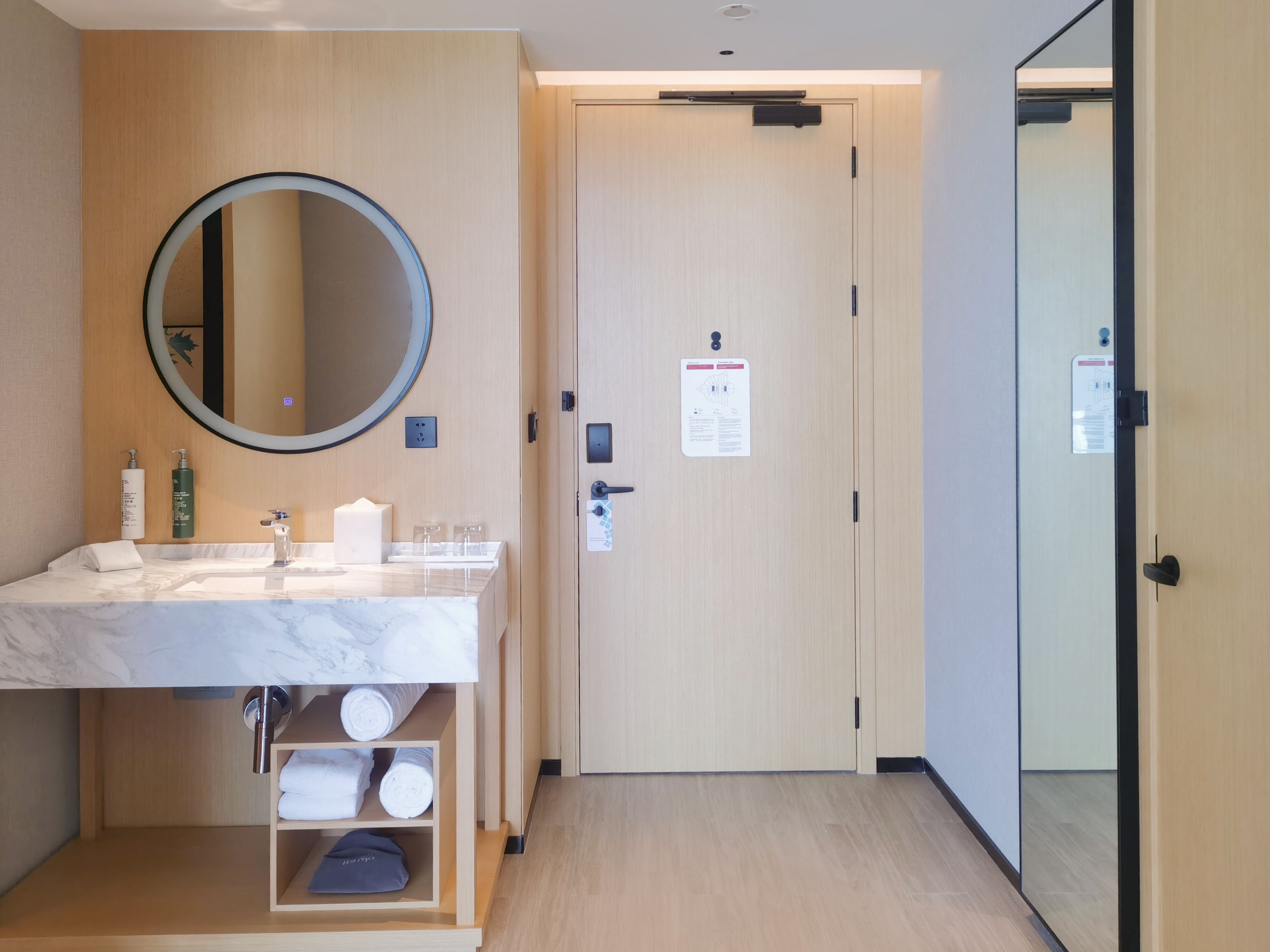 A hotel room with a marble sink, a round mirror, a wooden door, and a shelf with towels.