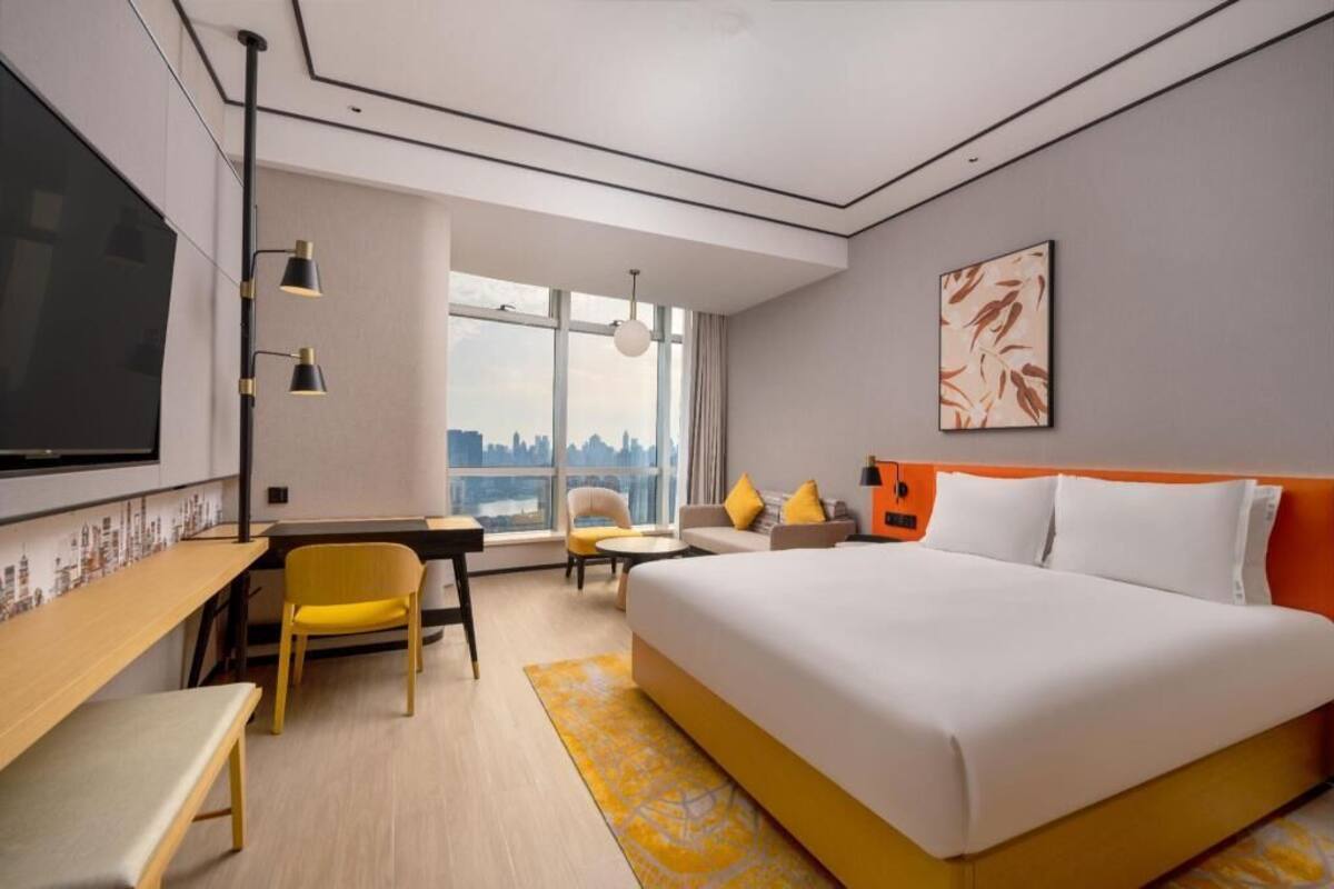 A hotel room with a bed, desk, chair, and a large window offering a city view.
