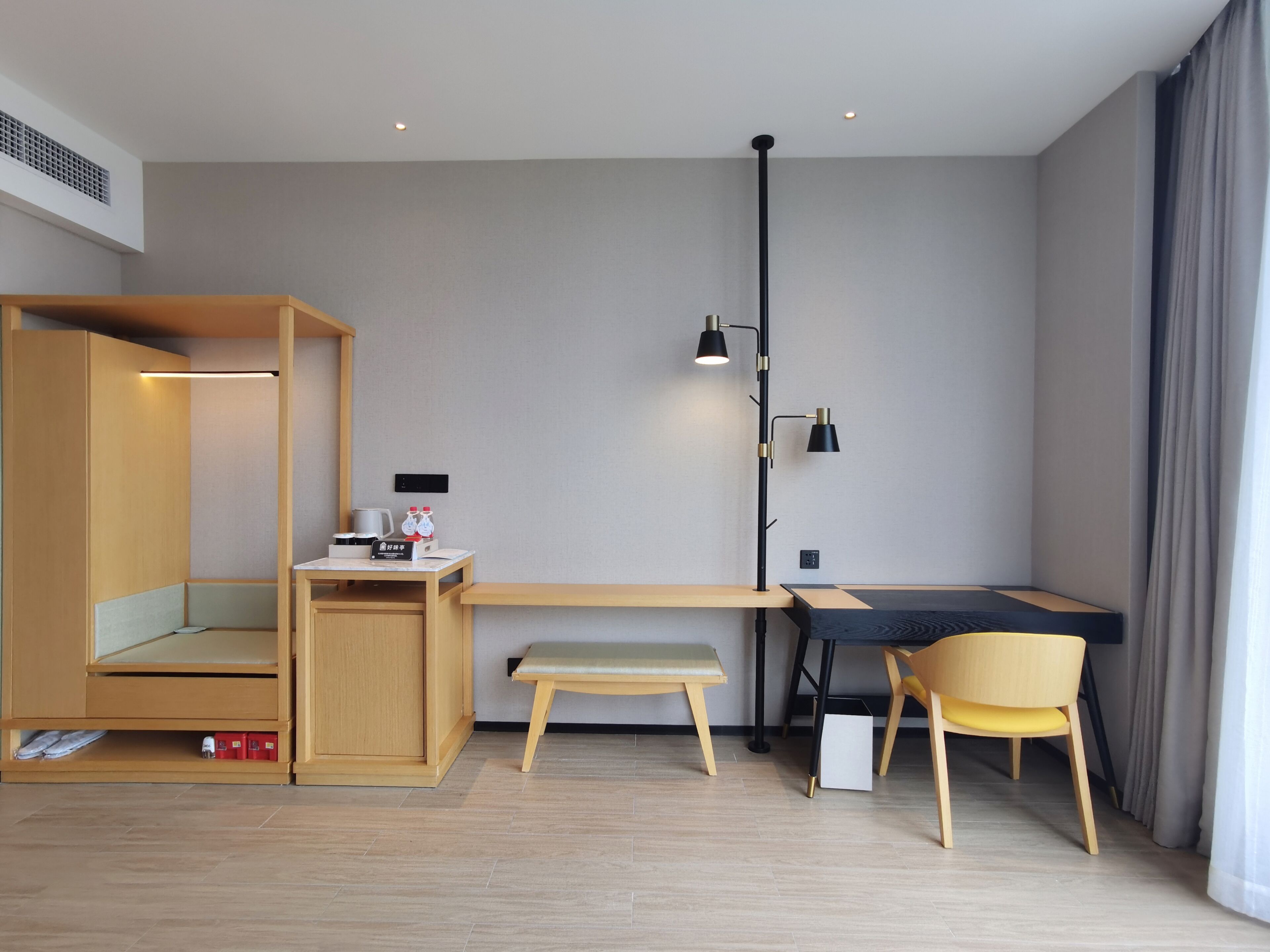 Una habitación de hotel moderna con un escritorio de madera, un banco y una silla, una lámpara de pie y una mesita con un hervidor y botellas.