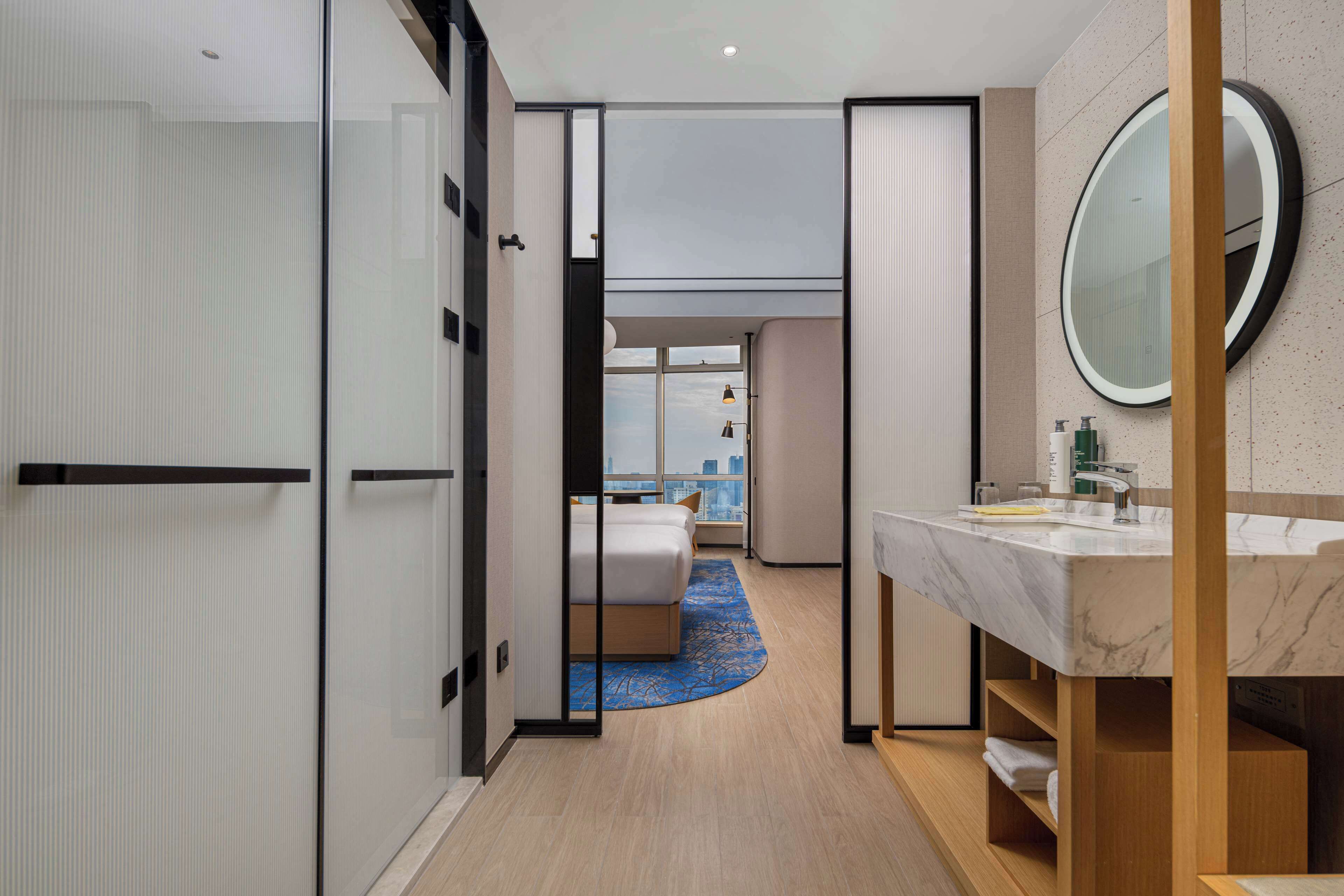 A modern hotel room with a large bed, a bathroom with a marble sink, and a view of the city through a large window.