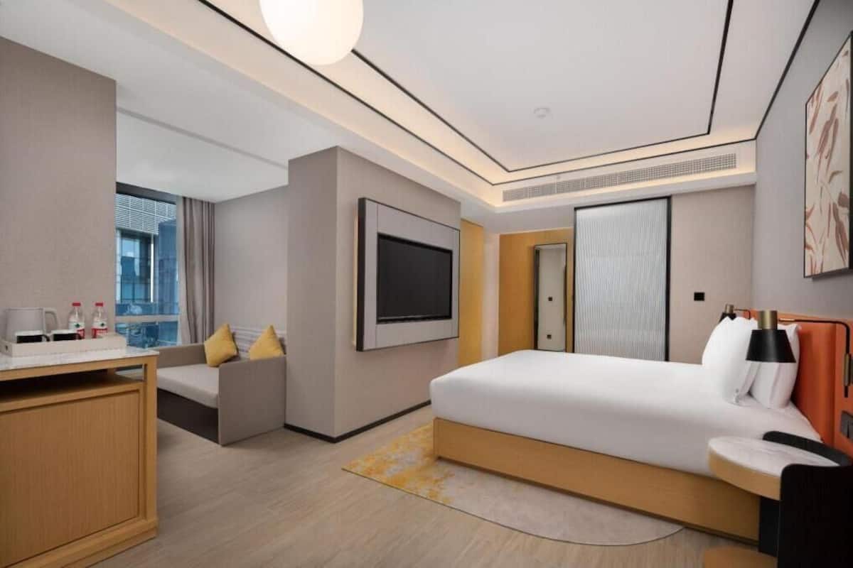 A modern hotel room with a bed, a TV, a sofa, a desk, and a window.