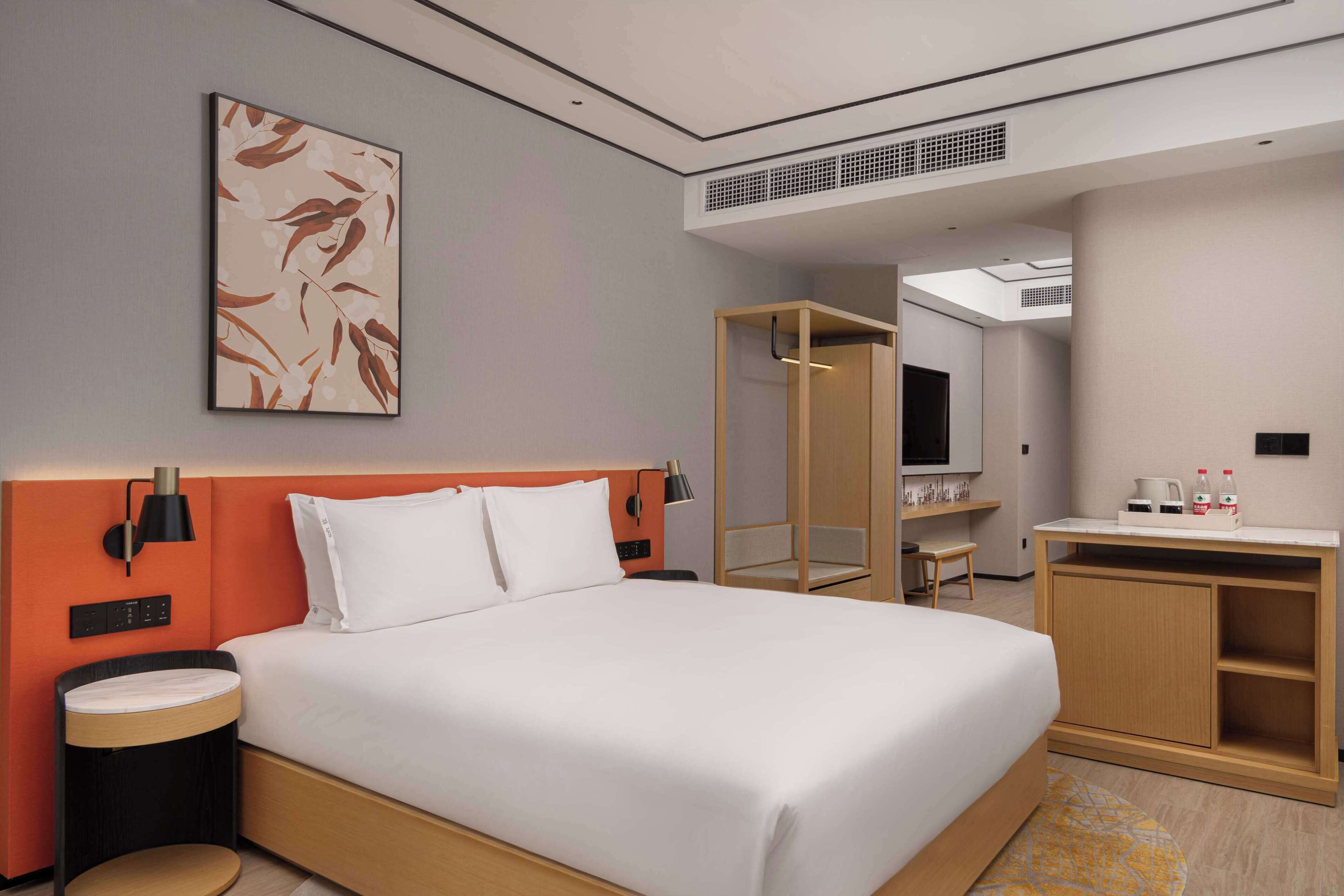 A modern hotel room with a large bed, a bedside table, a mirror, a wardrobe, and a small desk with a chair.