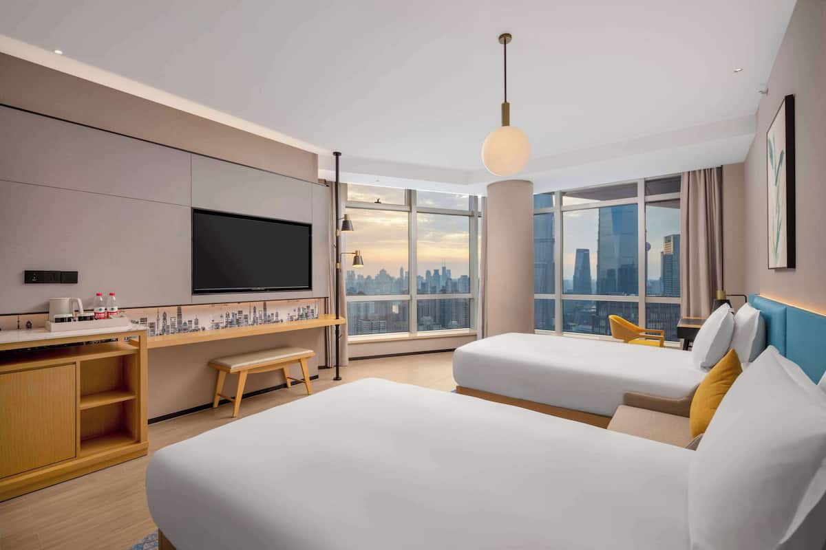 A modern hotel room with a large bed, a desk with a flat-screen TV, a coffee machine, and a city view.