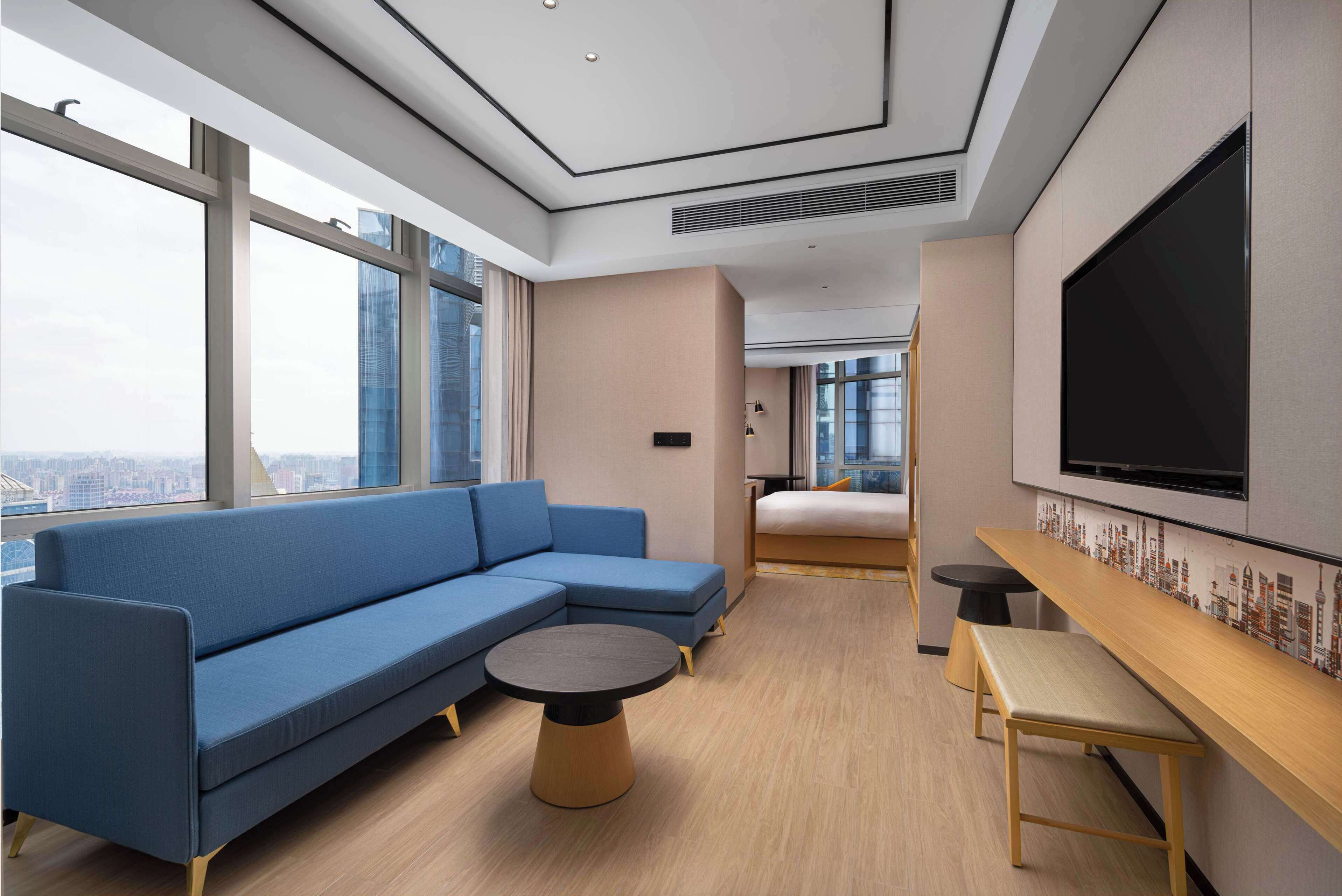 Une chambre d’hôtel moderne avec un grand canapé, une petite table ronde, un banc et un grand écran de télévision.