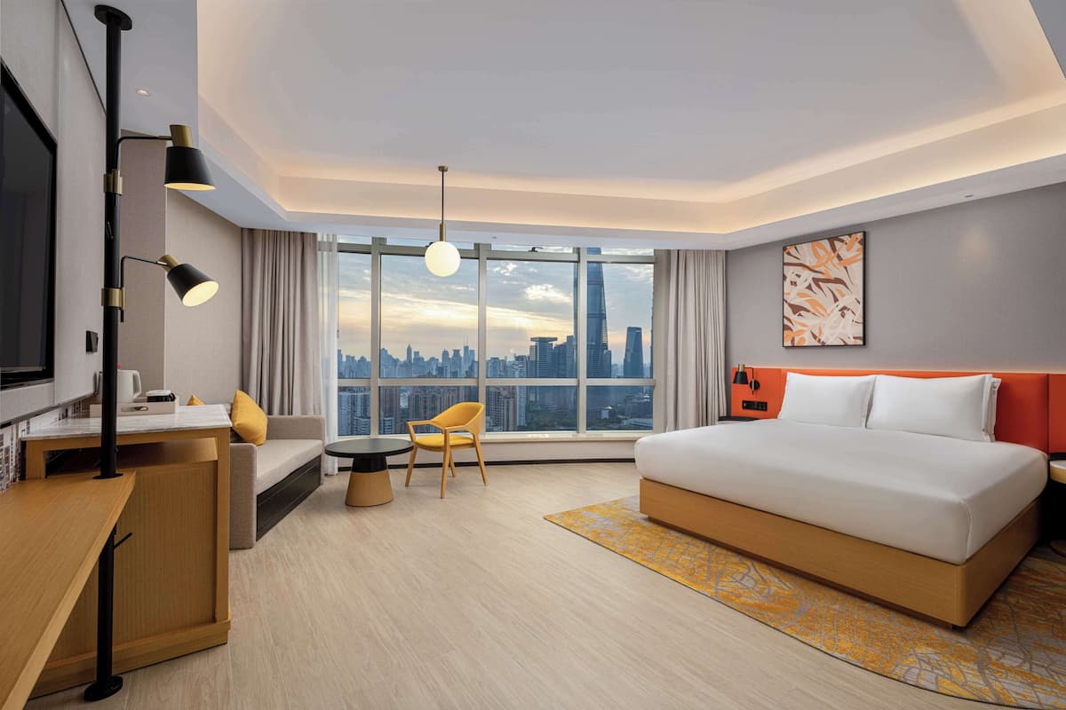 A modern hotel room with a large bed, a sofa, a chair, a desk, and a view of the city skyline through large windows.