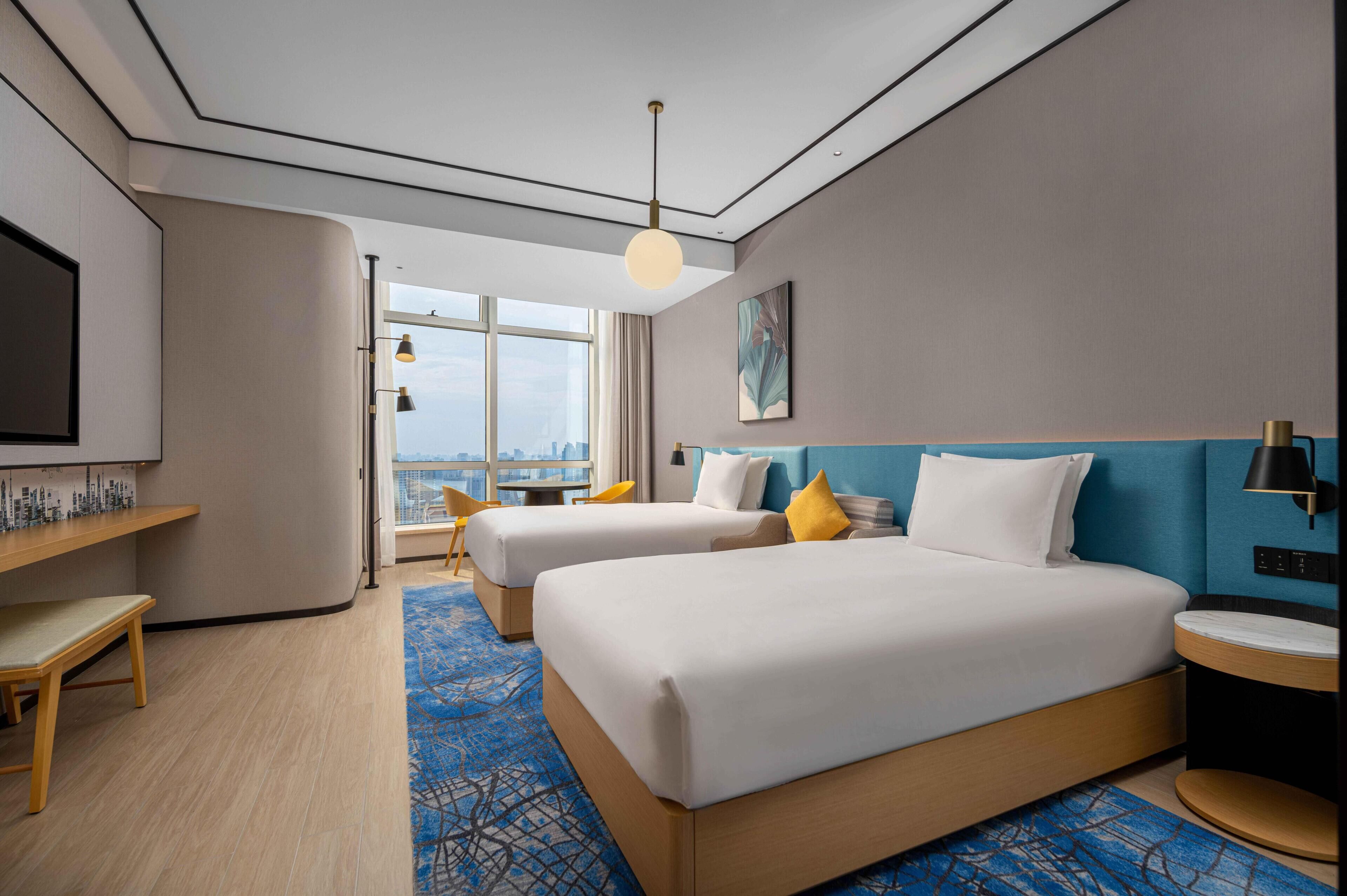A hotel room with two beds, a desk, a chair, a TV, and a large window with a city view.