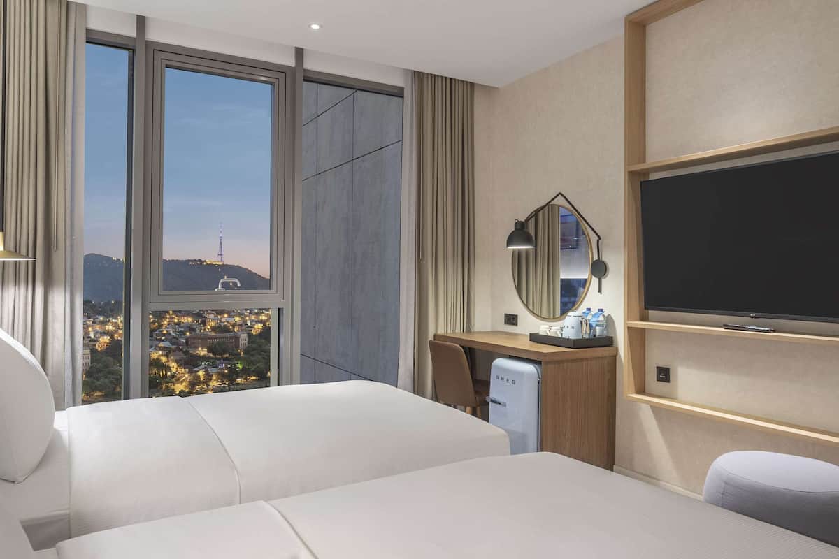 Hotelkamer met twee bedden, een bureau met computer, een televisie en een groot raam met uitzicht over de stad.